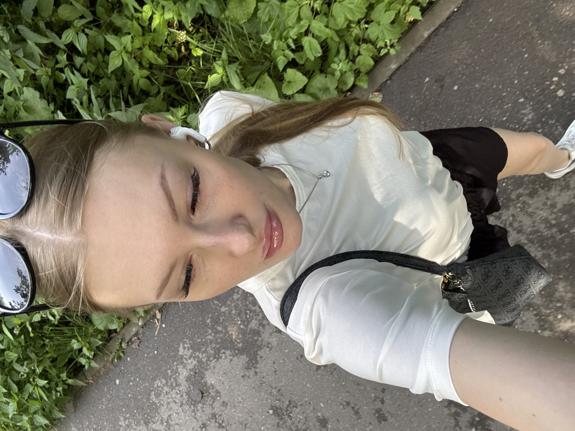A young woman with blonde hair, wearing white earbuds, a white t-shirt, and black shorts, is taking a selfie outdoors. She is looking down at the camera with her eyes partially closed, her lips slightly parted and glossy. She has on large, round sunglasses perched on her head. A small, dark-colored purse with a patterned design and a gold chain strap is slung across her body. The background consists of lush green foliage on the left and a paved pathway on the right. The scene suggests it is daytime, likely sunny, given the bright lighting and shadows. The setting appears to be a park or a natural area with a path for walking or jogging. The woman's expression could be interpreted as relaxed or playful. There is no visible text in the image. The location context is Perm, Russia.