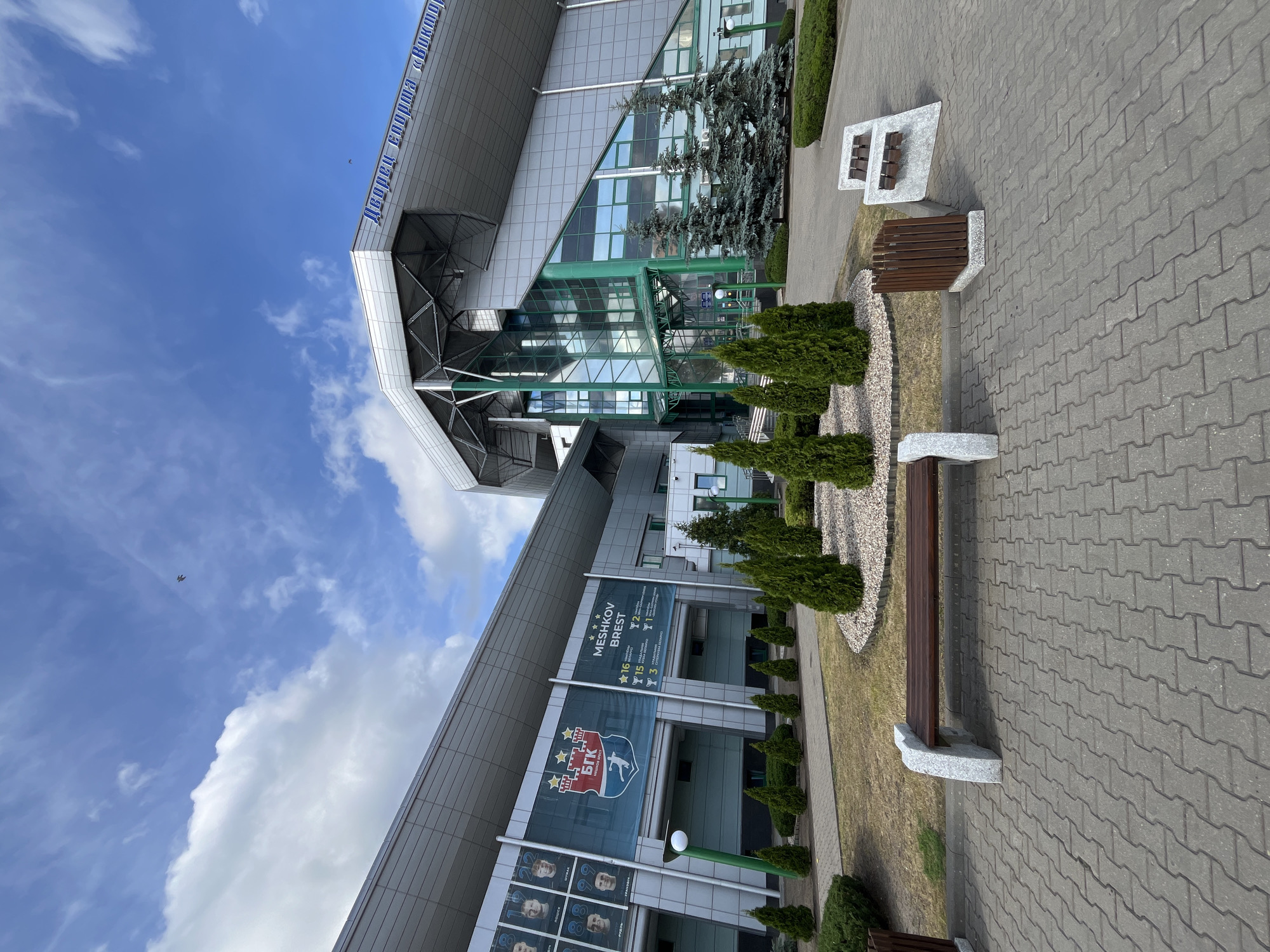 The image shows the exterior of a modern sports arena with a grey, metallic facade and a large glass entrance. The arena is named "Meshkov Brest" as indicated by a banner featuring the team's logo and some achievements. In front of the arena, there is a paved walkway with several benches and a waste bin. A small landscaped area with manicured evergreen bushes and trees is situated between the walkway and the building. The sky above is blue with scattered white clouds, and a single bird is visible flying in the distance. The overall scene suggests an outdoor area in front of a sports facility on a clear day.