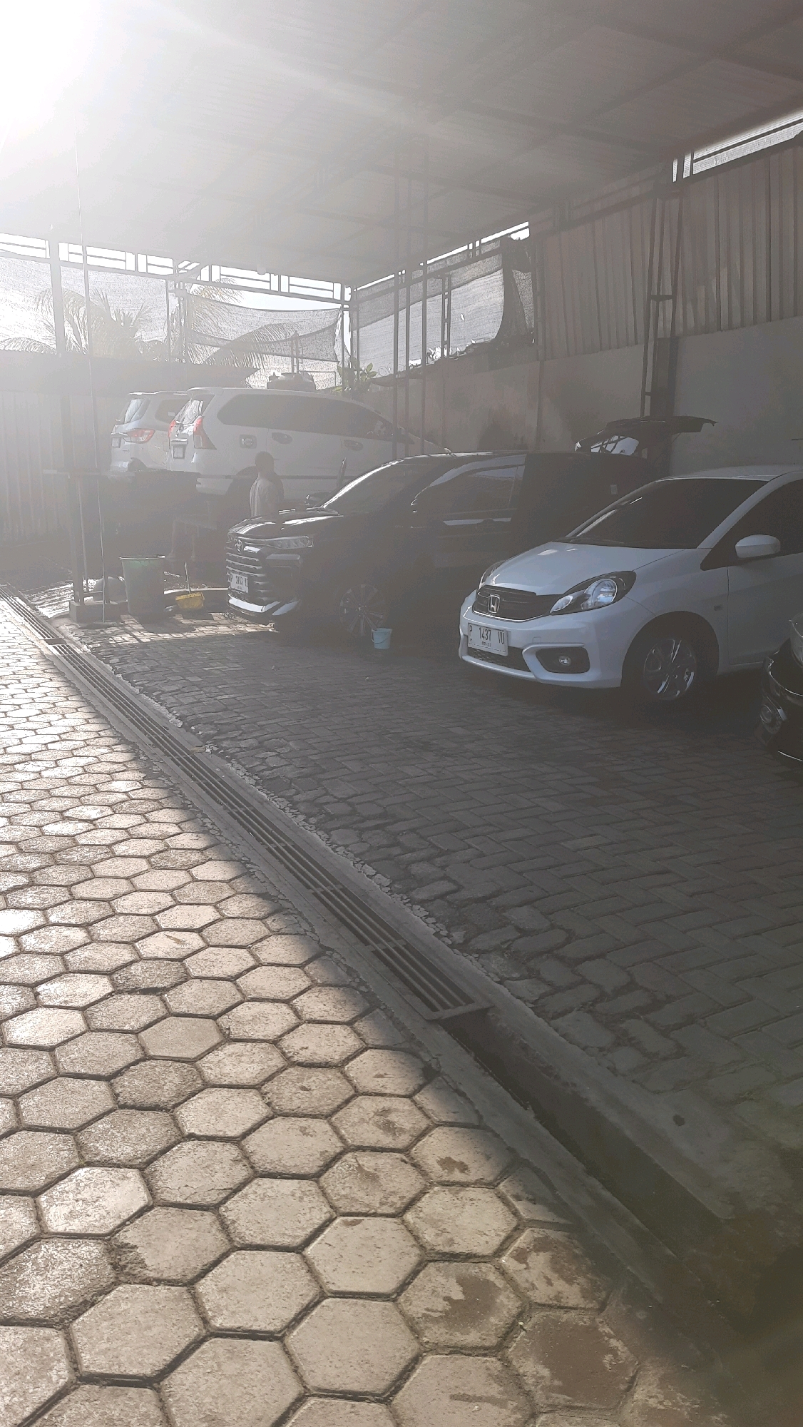 A car wash in Jajag, Indonesia, is depicted under a covered shelter. Several cars are parked in a row on a paved surface made of interlocking hexagonal stones and rectangular bricks. The sun is shining brightly, creating a hazy effect and casting shadows on the ground. In the background, palm trees are visible beyond the shelter. A person can be seen standing near a green bucket and a yellow mop, suggesting they are involved in the car washing process. The scene appears to be a typical day at a car wash, with vehicles waiting to be cleaned.