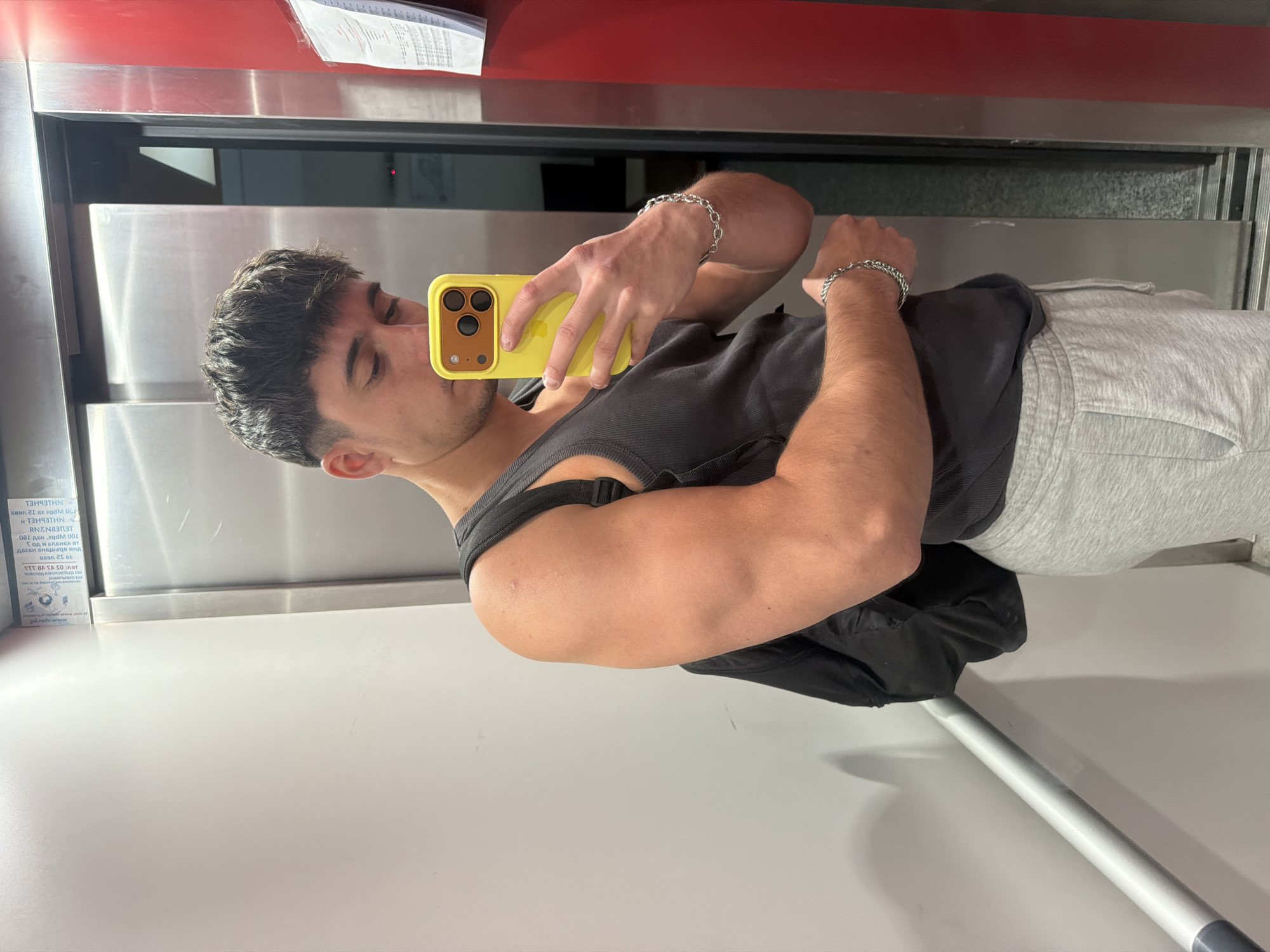 A young man is taking a selfie in what appears to be an elevator. He is wearing a dark tank top, light grey sweatpants, and silver chain bracelets on both wrists. He is holding a yellow phone with a brown camera area. The background shows the interior of the elevator with stainless steel walls and a red panel at the top. A product label with indecipherable text is affixed to the left wall. The scene appears to be indoors in an urban setting. There is no visual information to determine the location specifically within Sofia, Bulgaria.