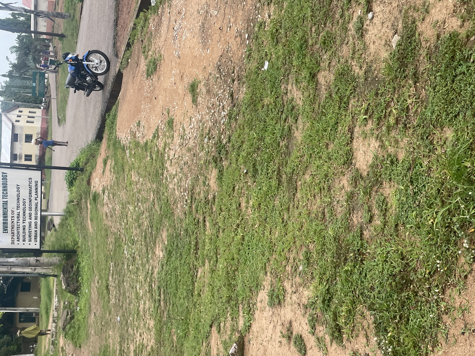 The image depicts an outdoor scene, likely on the grounds of an educational institution in Lokoja, Nigeria. In the foreground, a patch of dry, sandy soil is interspersed with clumps of green grass. Further back, a paved road curves into the scene. To the left of the road, a woman in a yellow veil walks away from the camera, carrying a round object on her head. Behind her, a light-colored building with visible windows stands. A signpost is visible, with text that appears to list departments of environmental technology.
To the right of the road, a blue motorcycle is parked. Beyond the motorcycle, a building with a yellow facade and white trim is visible, along with more trees and vegetation. In the middle ground, several people can be seen walking, including a young boy in a blue shirt and dark shorts. The overall impression is that of a university campus or similar educational facility, with a mix of buildings, roads, and natural elements.