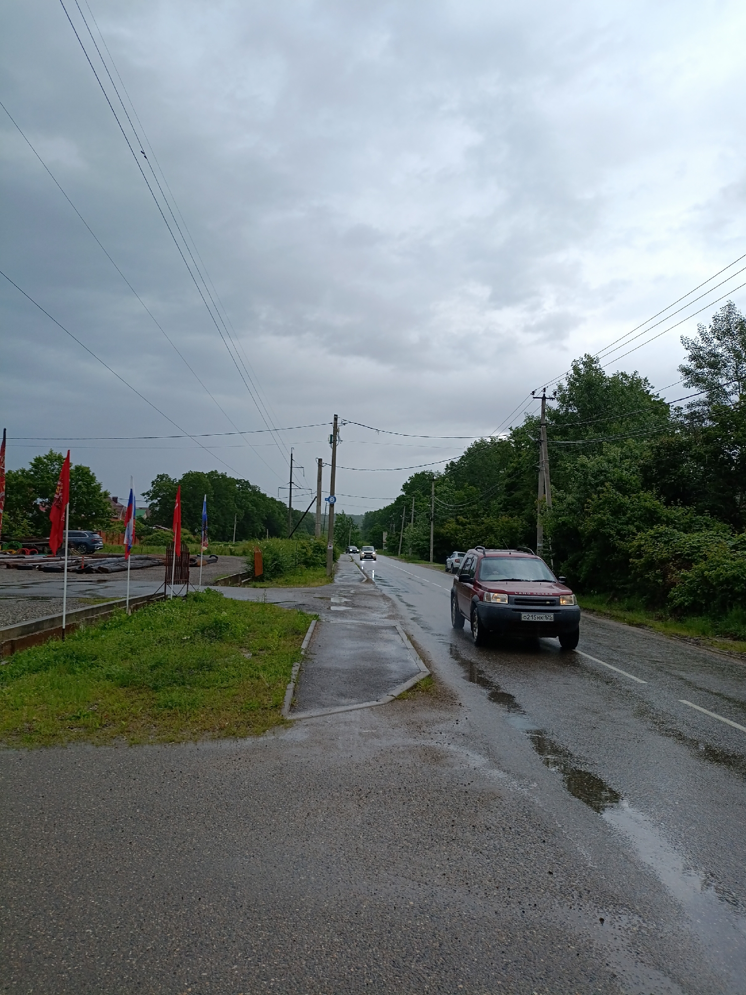 The image shows a wet road in Goryachiy Klyuch, Russia, under a cloudy sky. A red Land Rover is driving on the right side of the road, heading towards the viewer. On the left side of the road is a sidewalk and a grassy area with several flags, including the Russian flag. Utility poles and power lines run along the road. Trees line the road in the background. Other vehicles are visible further down the road. The road surface is wet, reflecting the overcast sky.