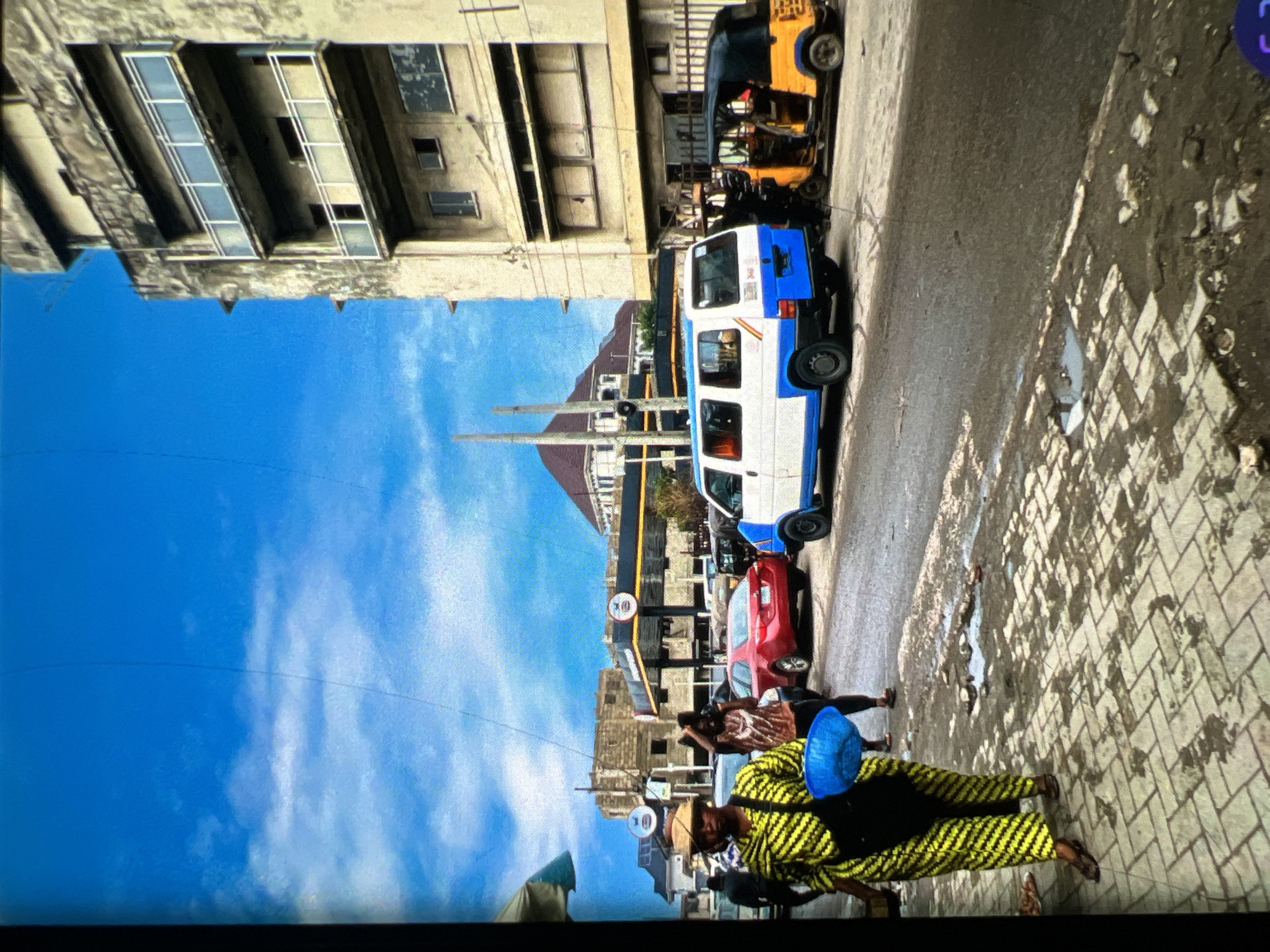 The image depicts a street scene in Lagos, Nigeria, under a bright blue sky with scattered clouds. In the foreground, a woman wearing a yellow and black patterned dress and a straw hat walks on a paved sidewalk, carrying a blue bowl and a black bag. To her right, a red car is parked on the wet asphalt street. Further down the street, a white and blue van is parked, and next to it, a yellow and black auto-rickshaw is visible. In the background, there is a multi-story building with balconies, and beyond that, a gas station with its canopy. Other people and vehicles are present in the mid-ground and background, suggesting a bustling urban environment. The street is marked by puddles and debris, indicating recent rainfall.