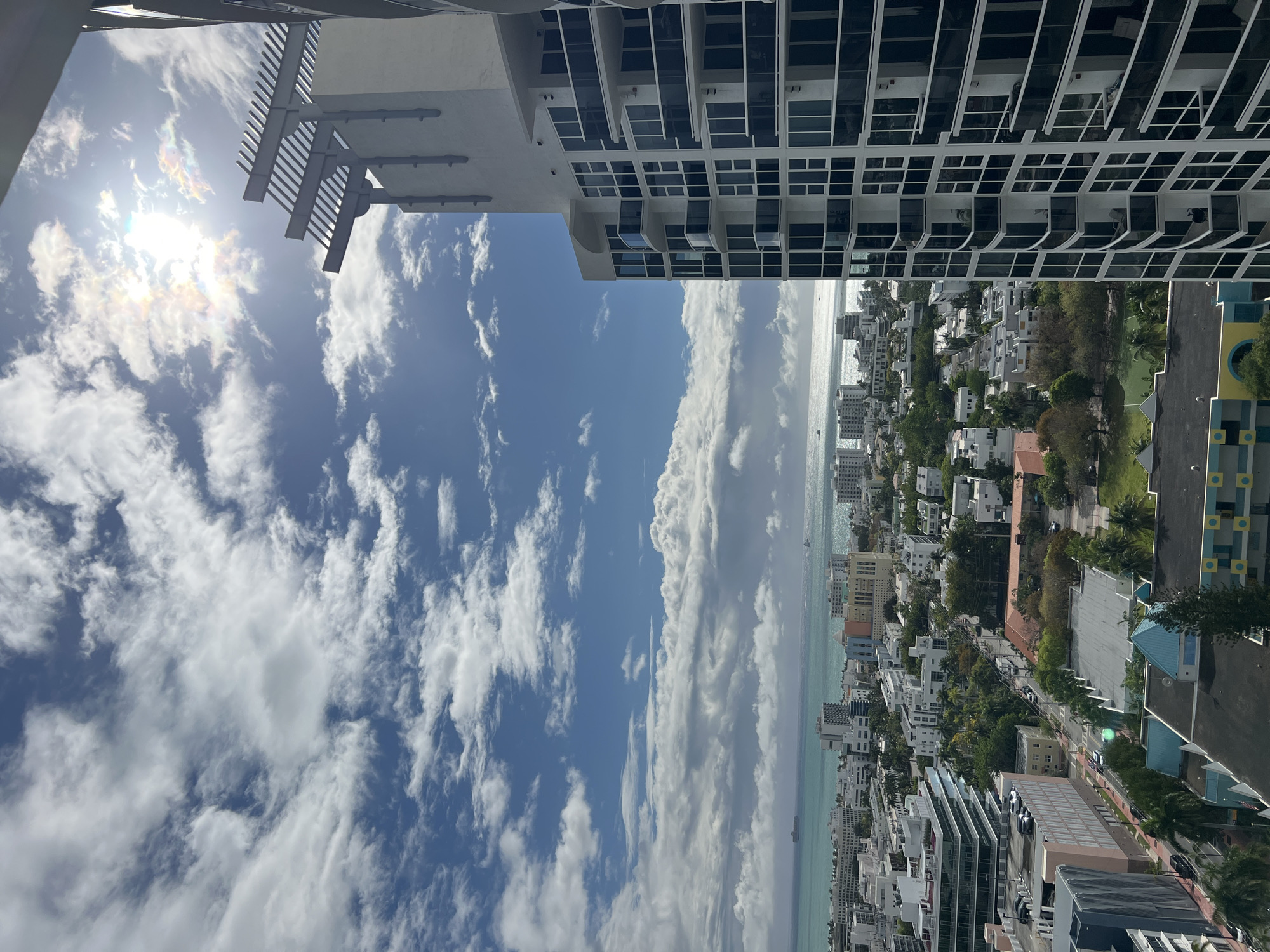 The image is a vertically oriented aerial shot of Miami Beach, Florida, United States. The perspective is from a high vantage point, likely a building or high-rise. The sky dominates a significant portion of the image, showcasing a blue sky filled with scattered white clouds. The sun is present but partially obscured by the clouds.
Below the sky is a coastal area with buildings. There are residential and commercial structures of varying sizes. Green trees are interspersed among the buildings. The ocean is visible in the distance.
The tops of buildings are seen in the upper part of the image. The architecture includes modern high-rise structures with balconies and repetitive window patterns. The balconies feature various objects.
There are no discernible people in the image. No specific actions or interactions are evident.