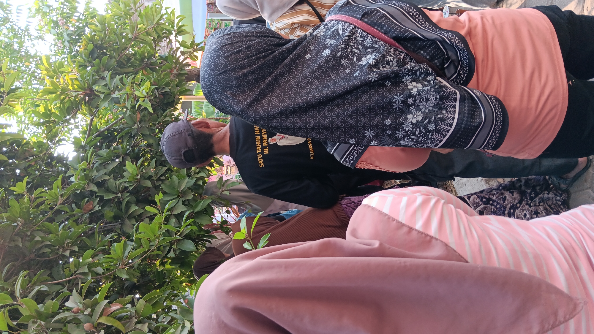 In Cerme, Indonesia, a group of people are gathered outdoors beneath a tree. The focus of the image is on the back of several individuals, most of whom are wearing headscarves or hijabs. One person in the center is wearing a black t-shirt with white text that reads "SATU TAHUN HAJI HI. PANIYE" and an image of a man in a white robe. This individual also wears a dark baseball cap. Another person in the foreground wears a pink and white striped shirt and a light pink hijab. To the right, a person wears a peach-colored shirt and a dark hijab with a floral pattern. The environment appears to be a casual outdoor setting, possibly a gathering or event, with the dappled sunlight filtering through the leaves of the tree.