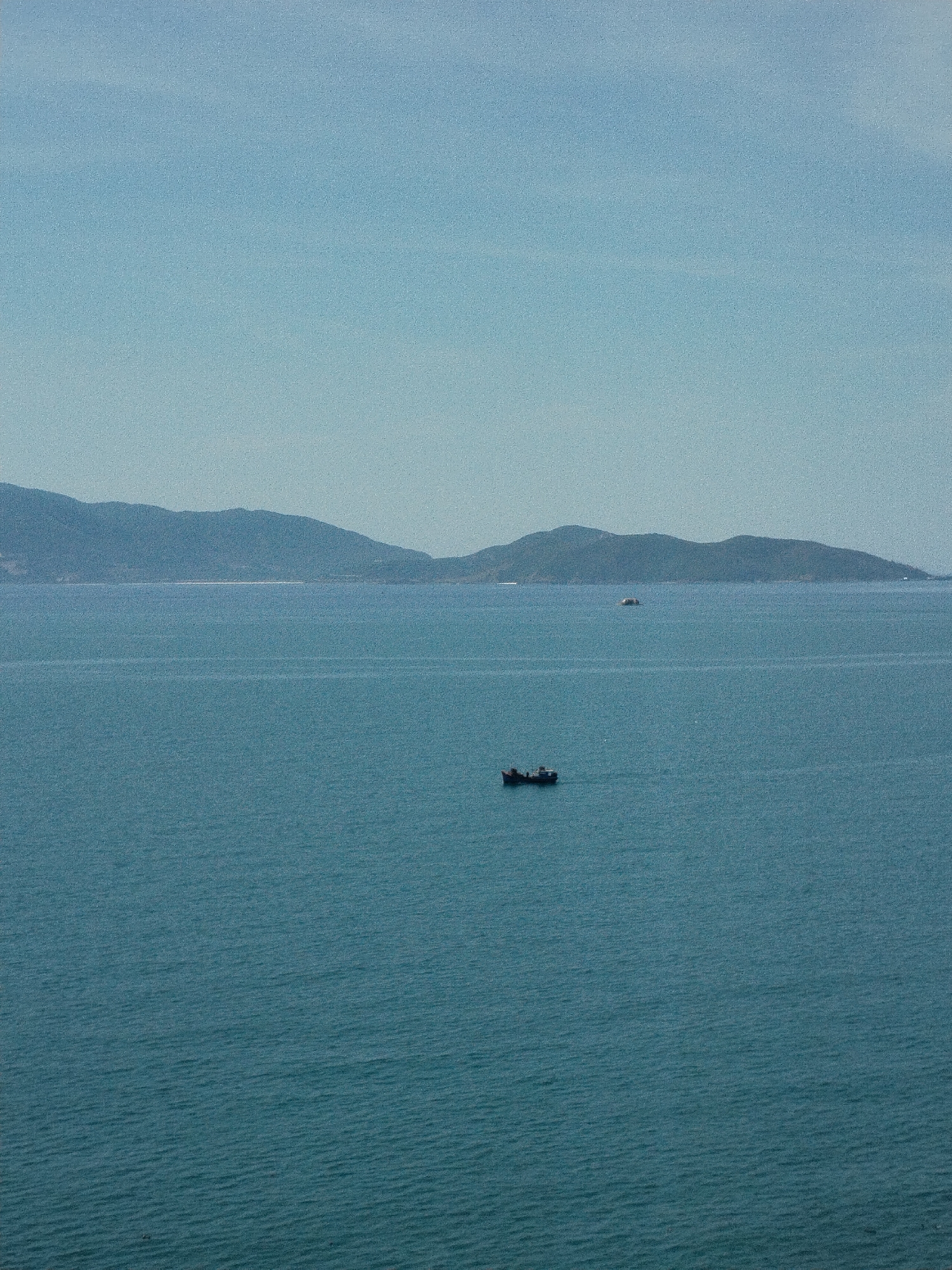 The image shows a body of water, likely the sea, with a small boat in the foreground and land visible in the distance, under a clear sky. The boat is located in the middle of the body of water with no visible people on board. The mountains appear to be in the far background, distant and muted in color. This appears to be in Bac Nha Trang Ward, Vietnam.