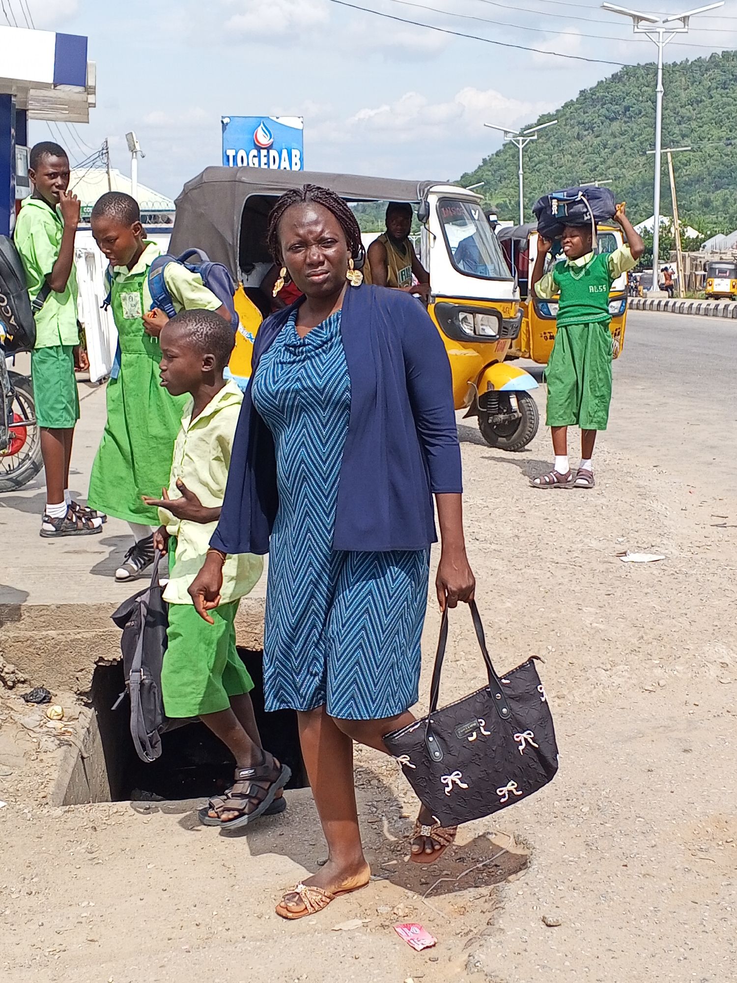 The image shows a group of people in what appears to be Zango-ndaji, Nigeria, based on signs in the background. A woman in a blue patterned dress and a navy blazer is standing in the foreground, holding a black purse with a white bow design. Behind her and to the left are three young children in school uniforms consisting of green and yellow attire, with backpacks. One child is stepping over a concrete drain. To the right, another child in a similar school uniform is carrying a bag on their head. In the background, there are yellow motorized tricycle taxis, one with a "TOGEDAB" sign on top, and a man sitting in the driver's seat. The environment includes a road, a petrol station, and a distant hilly landscape under a partly cloudy sky.