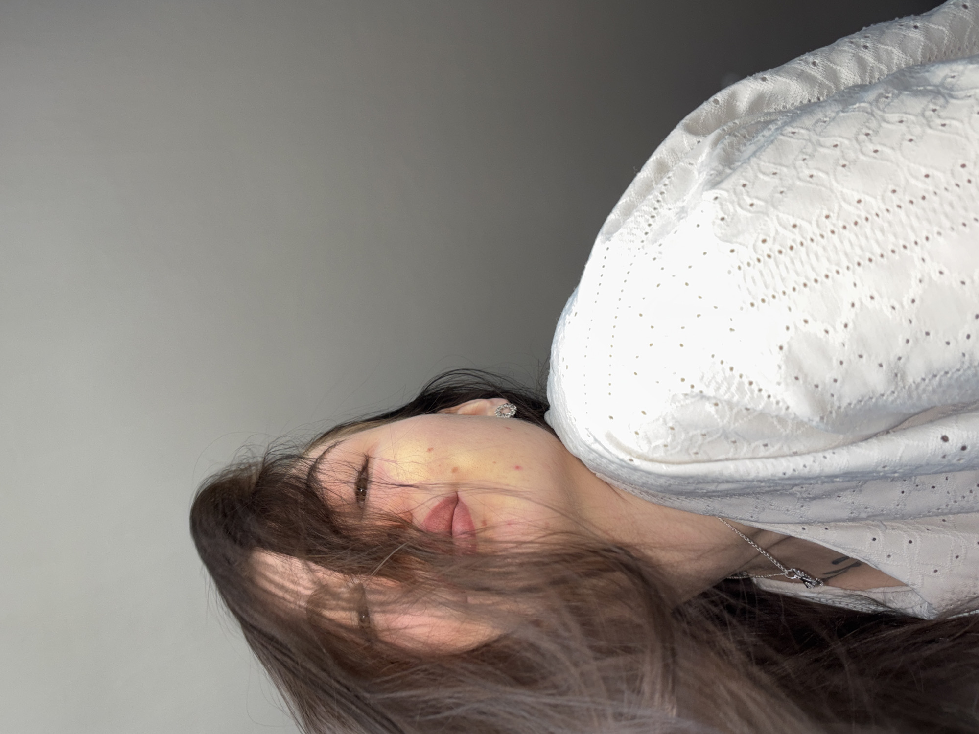 A woman with long, brown hair is lying down. Her face is partially obscured by her hair. She is wearing a white, eyelet-style top and a silver necklace. She has fair skin and a small earring. The background is a gradient of grey. The image does not contain enough information to determine the location in Perm, Russia.