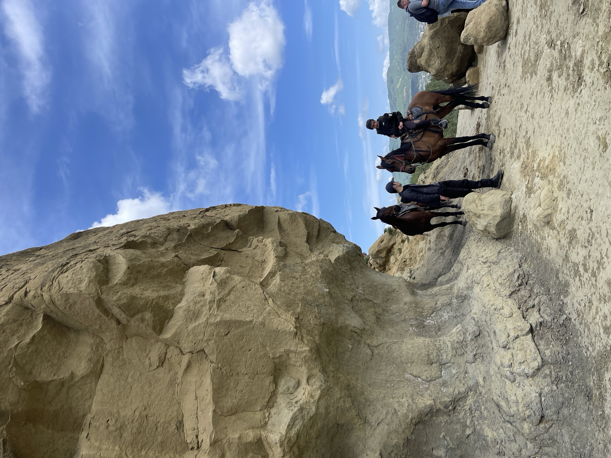The image shows a group of people on horseback in a rocky, mountainous environment, possibly in the vicinity of Мирный, Russia. The foreground is dominated by a large, light-brown rock formation. Three horses with riders are positioned on a flatter, rocky surface to the right of the frame. One person is standing on the ground next to a horse, while others are sitting on the rocks at the very top of the picture. In the background, a valley and distant hills are visible under a blue sky with scattered clouds.