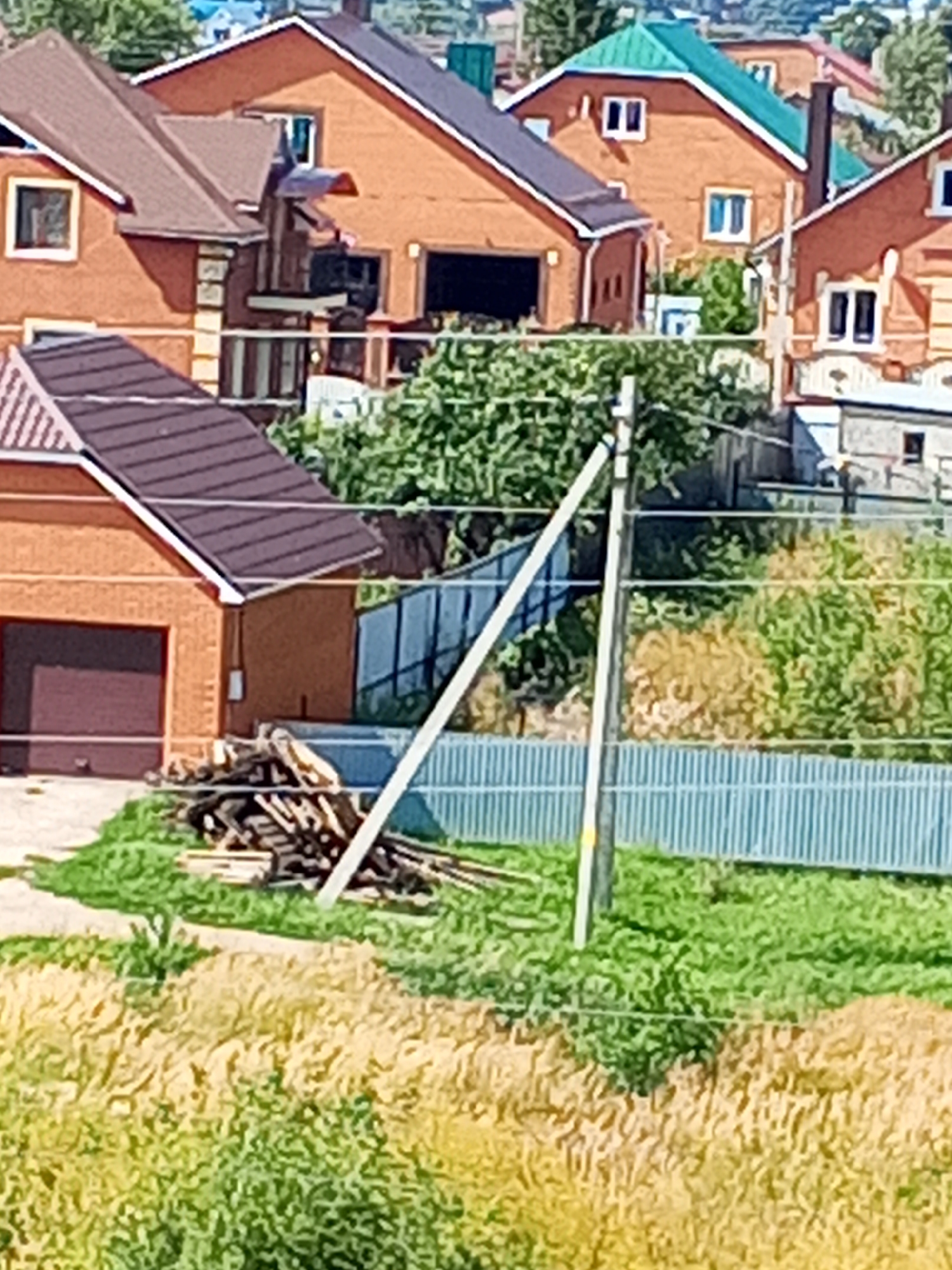 The image depicts a residential area with several brick houses under a clear sky. In the foreground, there is a pile of lumber next to a garage door. A light blue fence runs horizontally across the middle ground, with a light-colored utility pole behind it. The houses are of various sizes and colors, with red brick being a prominent material. Some houses have green roofs, while others have brown or gray ones. The overall environment appears to be a semi-urban or suburban setting, with vegetation and trees visible around the houses. The specific location is in Chistopol, Russia.