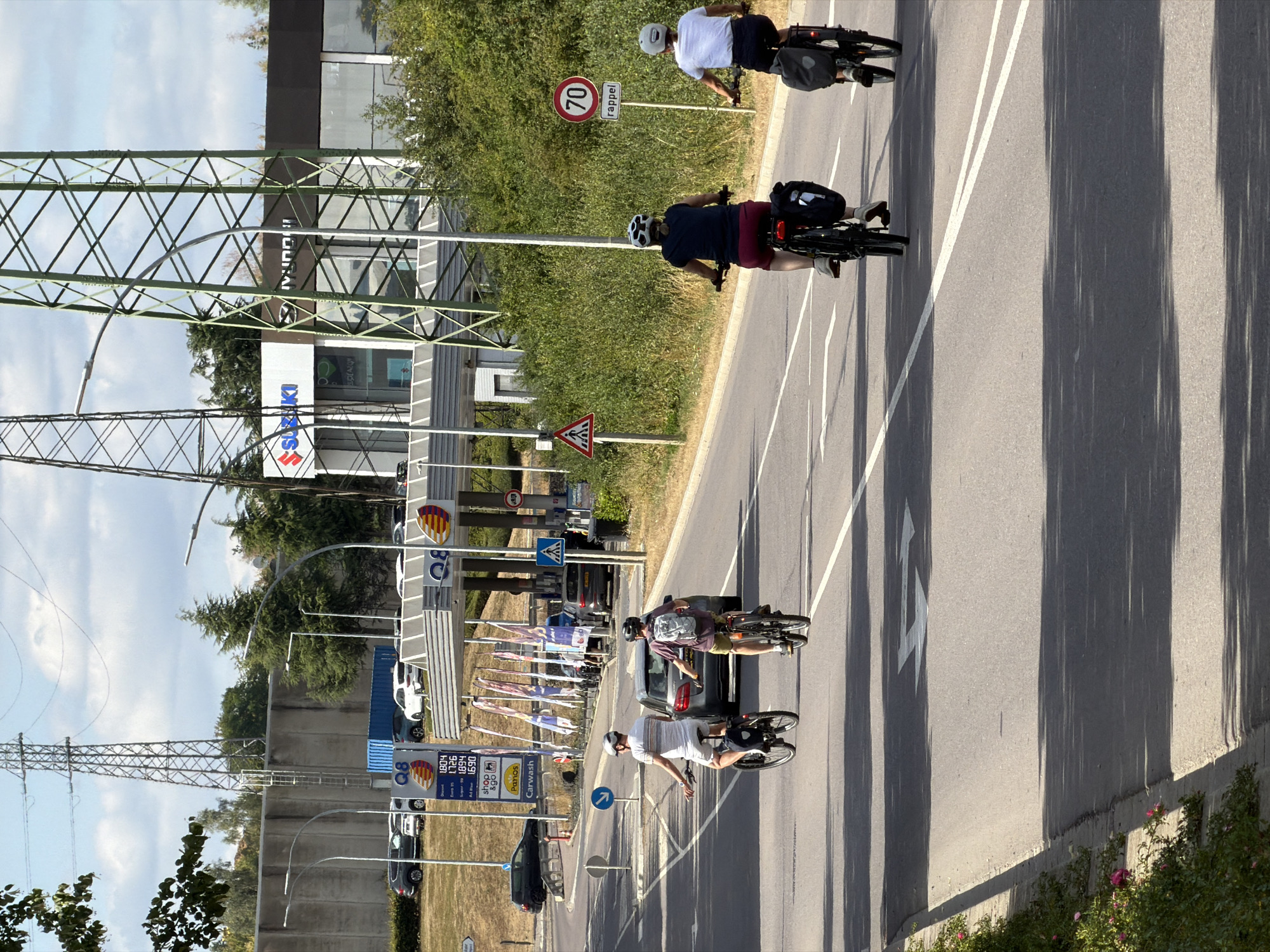 In Esch-sur-Alzette, Luxembourg, on a sunny day, a paved road stretches out under a partly cloudy sky. Tall electrical towers loom in the background, with one featuring a green metal lattice structure and another a silver lattice. A series of buildings are visible, including a Q8 gas station with multiple pumps and prominent Q8 branding, and other structures with SUZUKI and HYUNDAI signage. Cars are parked and driving on the road, and a grey Mercedes sedan is in the foreground.
Several individuals are engaged in activities: two people are cycling on the road, wearing helmets and riding bicycles equipped with panniers. Another two individuals are also cycling, one on a black bicycle with a backpack and the other on a black bicycle with panniers. A pedestrian crossing sign is visible, as well as a speed limit sign indicating 70. Lush green trees and bushes line the roadside, adding to the natural scenery. The road has clear lane markings, including white lines and directional arrows.