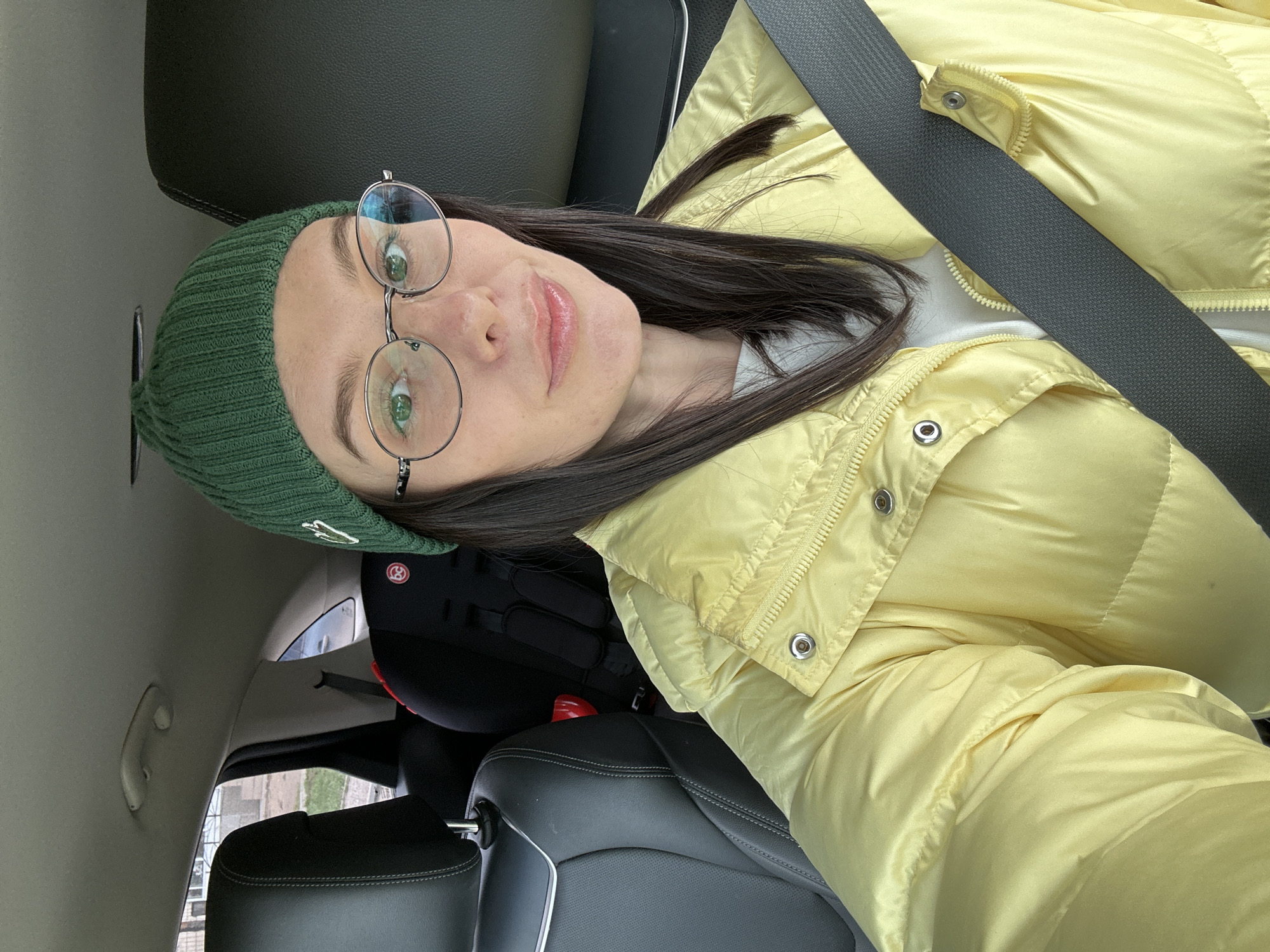 A woman is sitting in the driver's seat of a car, wearing a yellow puffer jacket, a green knit hat with a logo, and round eyeglasses. She is wearing a seatbelt. A child's car seat is visible behind her in the back seat. The car is parked and the surrounding environment is visible through the car windows, including buildings and greenery. The location may be Chaikovsky, Russia.