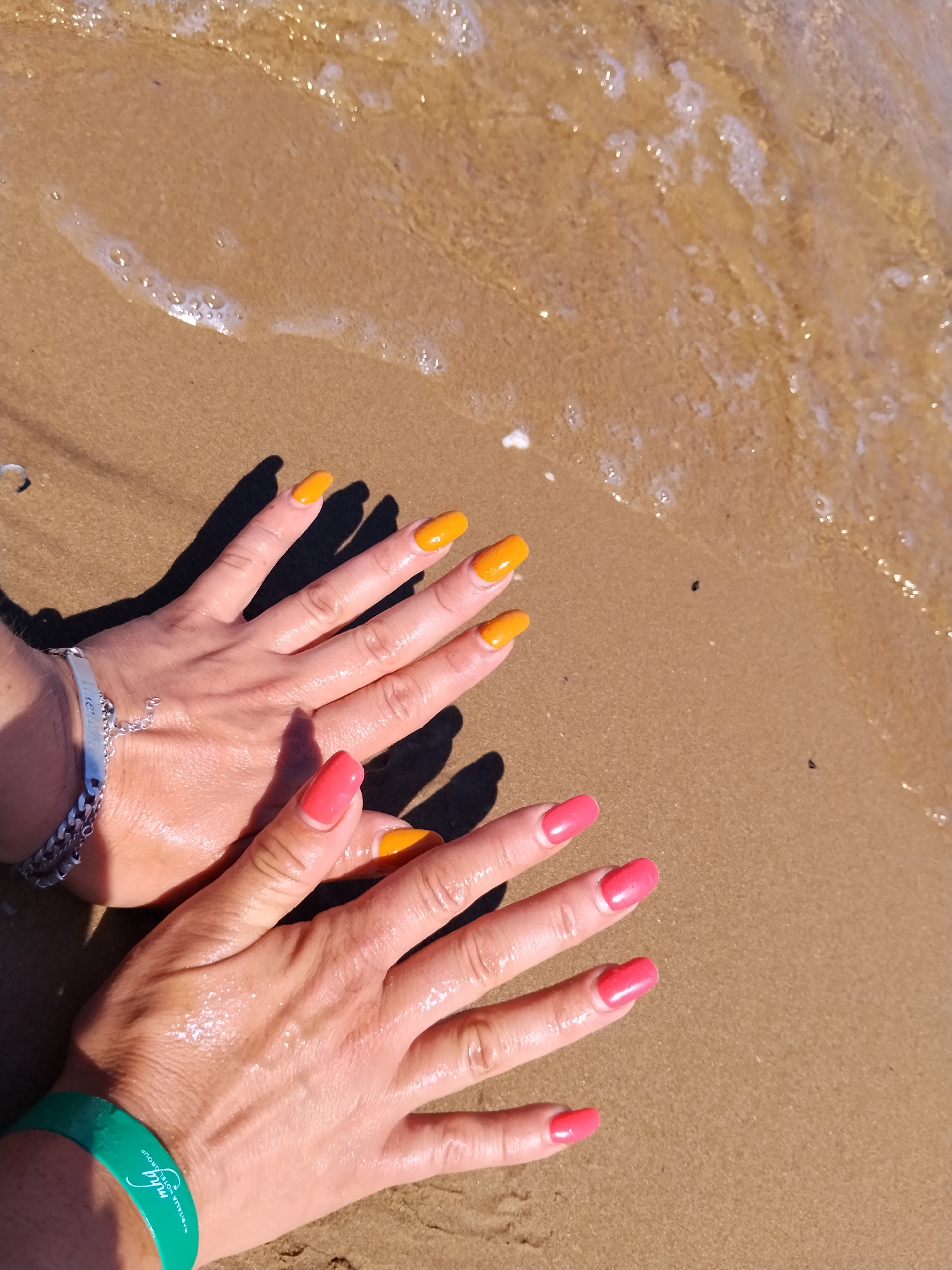 This is a high-angle, medium shot photograph of two hands with painted fingernails on a sandy beach. The hands are resting on wet sand, with the edge of the water lapping at the sand near the upper right quadrant of the image. The fingernails are painted in two different colors: bright orange on the upper hand and a vibrant pink on the lower hand. The upper hand has a silver bracelet on the wrist, and the lower hand has a green wristband with white lettering that appears to read "Murgia Hotel Group". The lighting suggests it is daytime, with distinct shadows cast by the hands. The environment is a beach, with wet sand and the gentle waves of the sea. The location is the beach in Peschici, Italy.