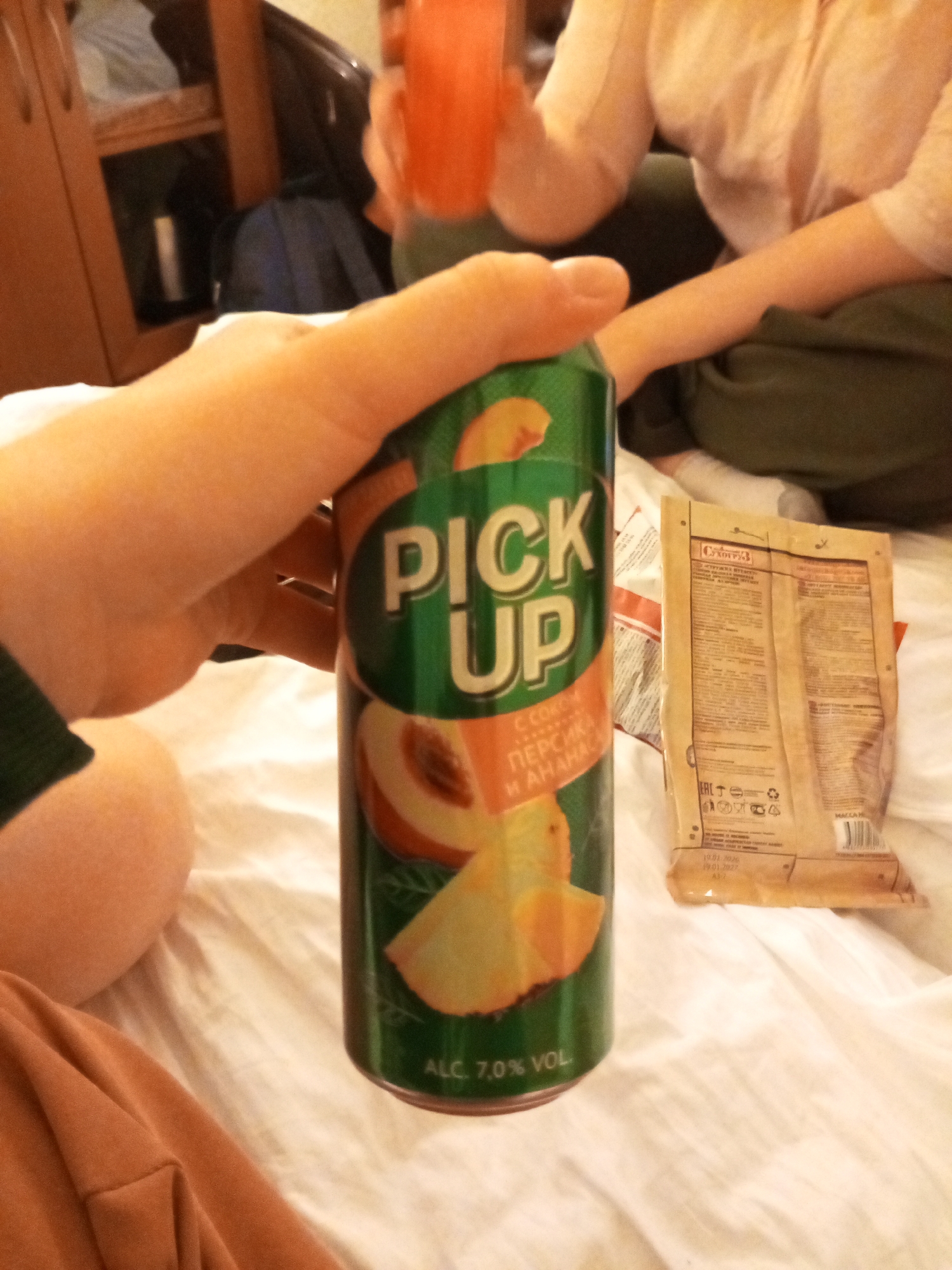 The image depicts a person holding a green can of "Pick Up" soda with peach and pineapple flavors, as indicated by the image on the can and the Cyrillic text "ПЕРСИКА И АНАНАС". The can is marked "ALC. 7.0% VOL." Also visible is a package of "Сухарики" (Croutons), a type of snack.
The setting appears to be indoors, possibly a bed, with a white sheet visible. Another person is seated in the background. There is a wooden cabinet or shelf in the upper left.