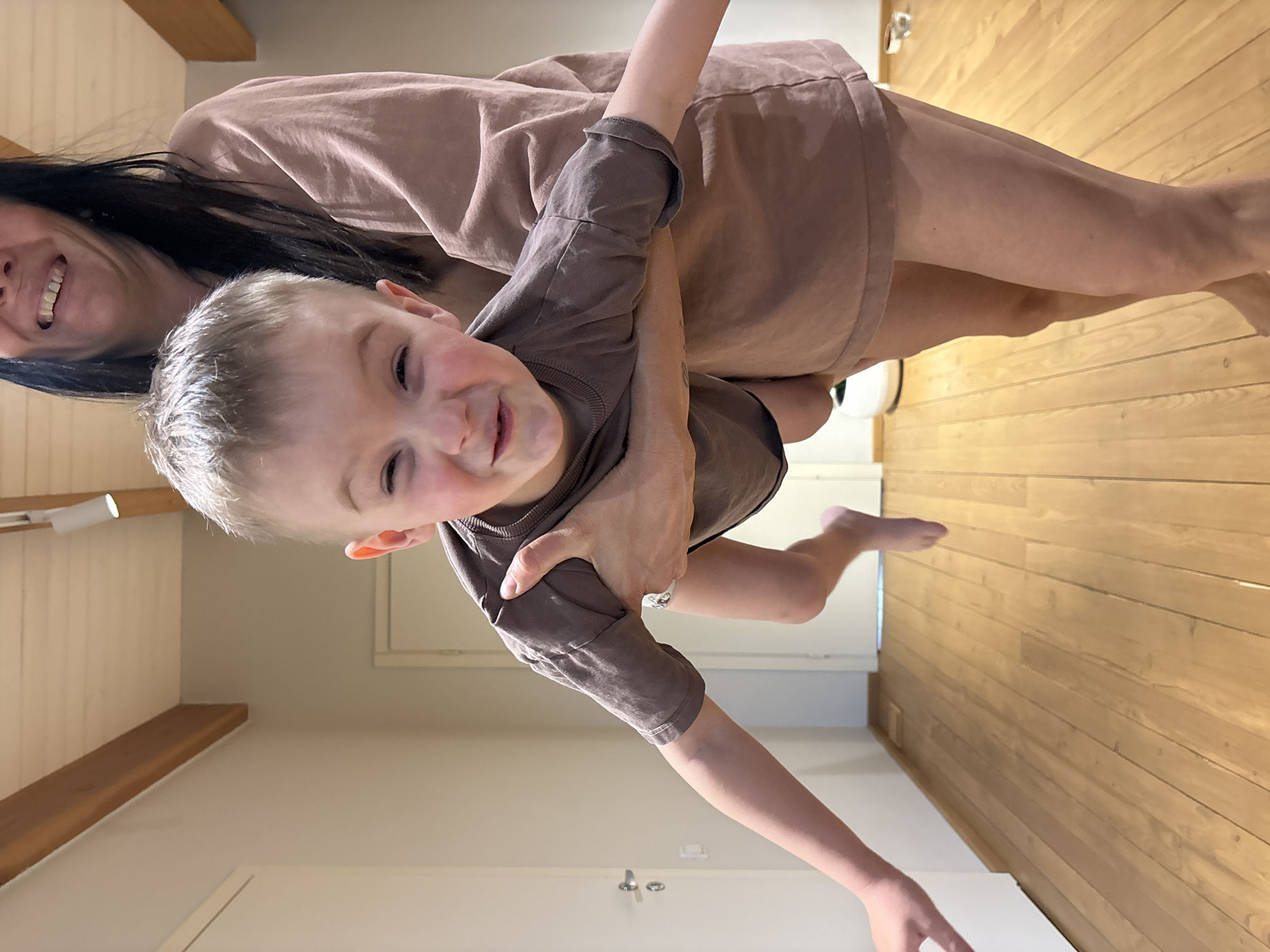 A woman is holding a young boy upside down in a hallway with wooden floors. The woman has long dark hair and is smiling. The boy is wearing a brown shirt and shorts, and also smiling. The hallway is brightly lit, with a white door on the left side of the frame. The location is inside a building, and is not related to the city of Chaikovsky, Russia.