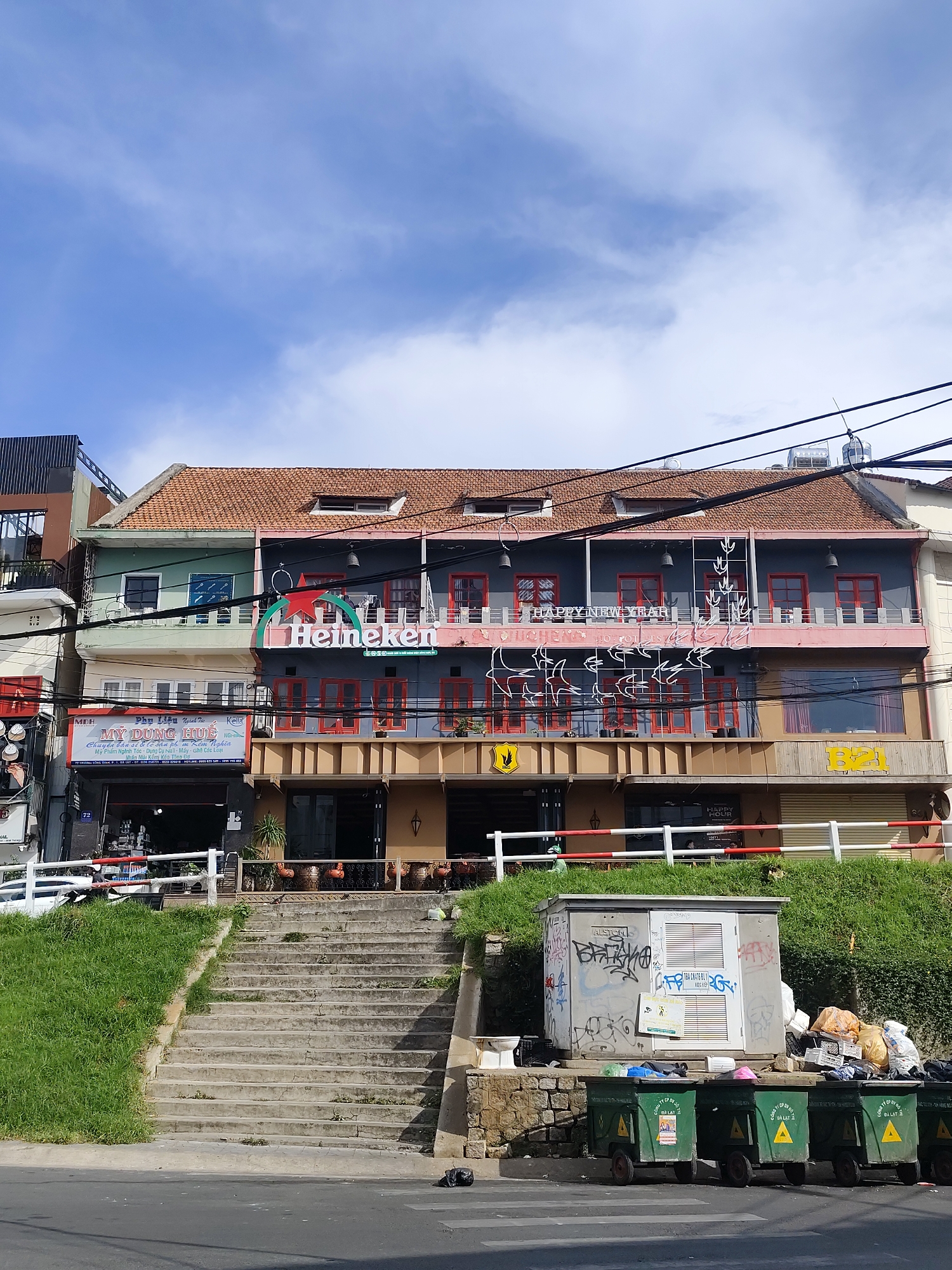 The image depicts a street scene in Da Lat, Vietnam, featuring a multi-story building with storefronts and a Heineken advertisement. A set of concrete stairs leads up to the building from the street, flanked by grassy slopes. In the foreground, several green trash bins are lined up next to a gray electrical box covered in graffiti. The building itself has a reddish-orange tiled roof and multiple levels with windows and balconies. The sky above is a bright blue with scattered white clouds. The overall impression is one of a commercial street with a mix of modern advertising and a slightly unkempt environment, indicated by the overflowing trash bins and graffiti.