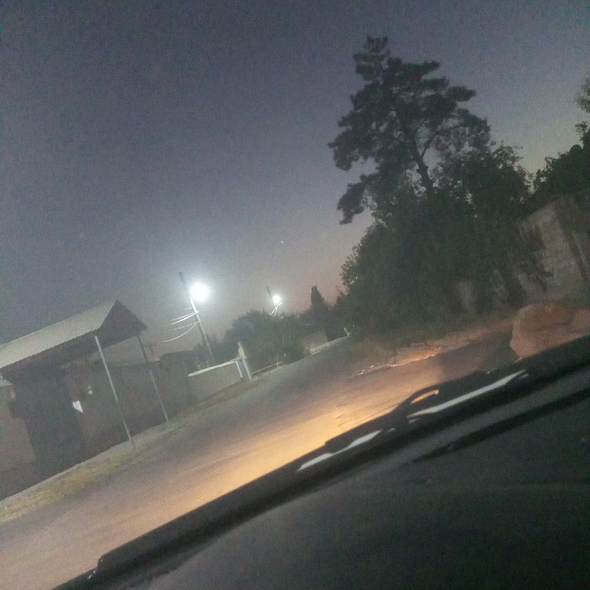 The image captures a dimly lit outdoor scene at what appears to be dusk or night, possibly in Chighirik, Uzbekistan. In the foreground, the edge of a vehicle's dashboard and windshield are visible, with the windshield wipers partially obscuring the view. The road ahead is illuminated by headlights, creating a bright yellow and white glow that reflects on the asphalt. To the left, a simple, covered structure with a sloped metal roof supported by posts is present, possibly a bus stop or a small shelter. A brightly lit street lamp stands further down the road, casting its light into the environment. The background features dense foliage and trees, with a prominent pine tree on the right side of the frame. A light-colored wall or fence is also visible along the right side of the road. The sky is a muted grey, suggesting the ambient light is low. There are no people clearly visible in the image.