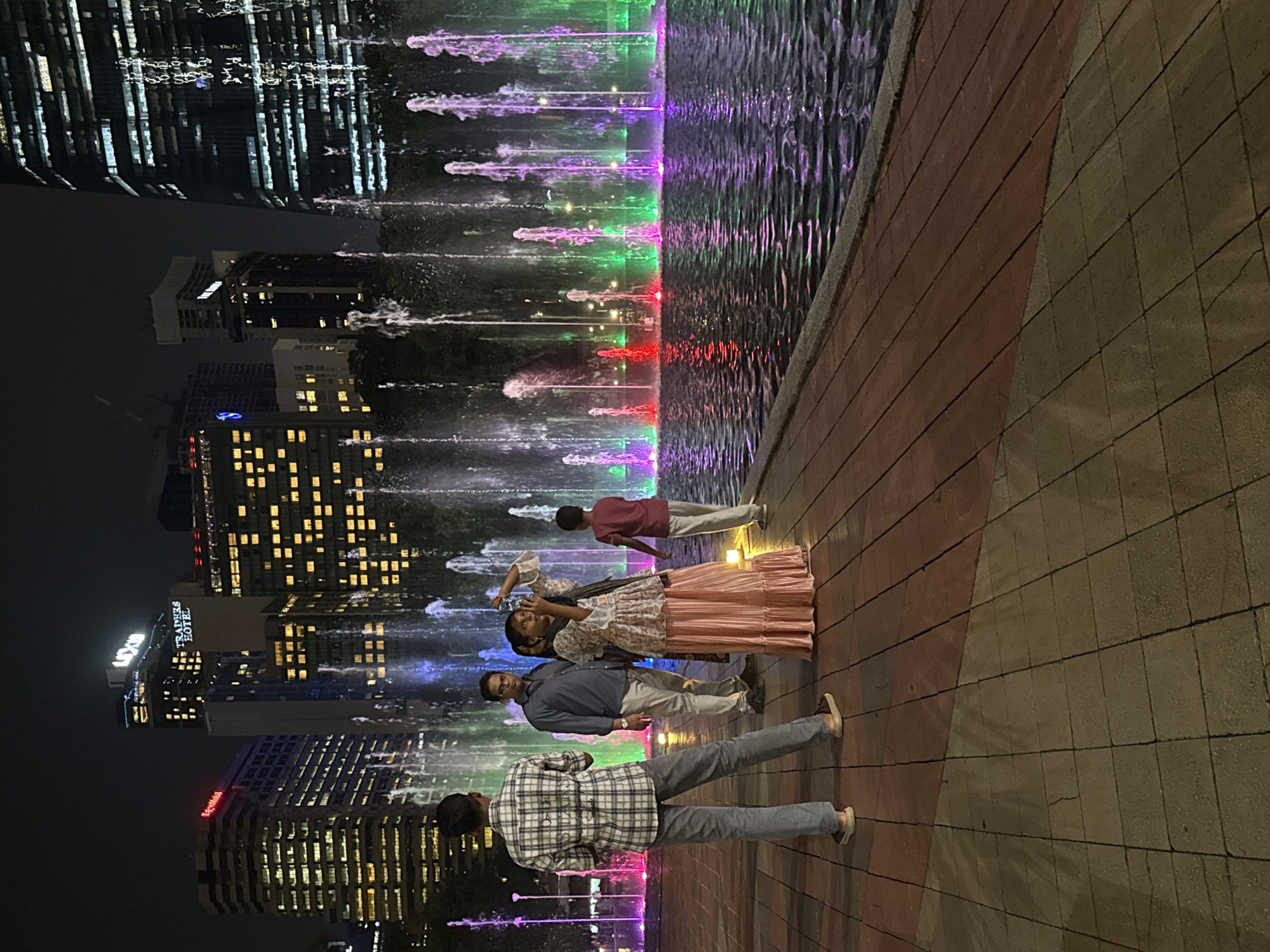 At night, a group of four people is standing in front of a colorful fountain at the KLCC Park in Kuala Lumpur, Malaysia, with the city skyline in the background. The fountain features illuminated water jets in various colors, including purple, blue, green, and red. The ground is paved with square bricks. The skyline consists of tall buildings with many lit windows. The first person, closest to the viewer, is wearing a plaid shirt and grey pants. The second person wears glasses and is dressed in a grey jacket and white pants. The third person is taking a photo with a phone; the person is wearing a floral shirt and a peach-colored skirt. The fourth person, standing slightly apart from the group, is wearing a red shirt and white pants.