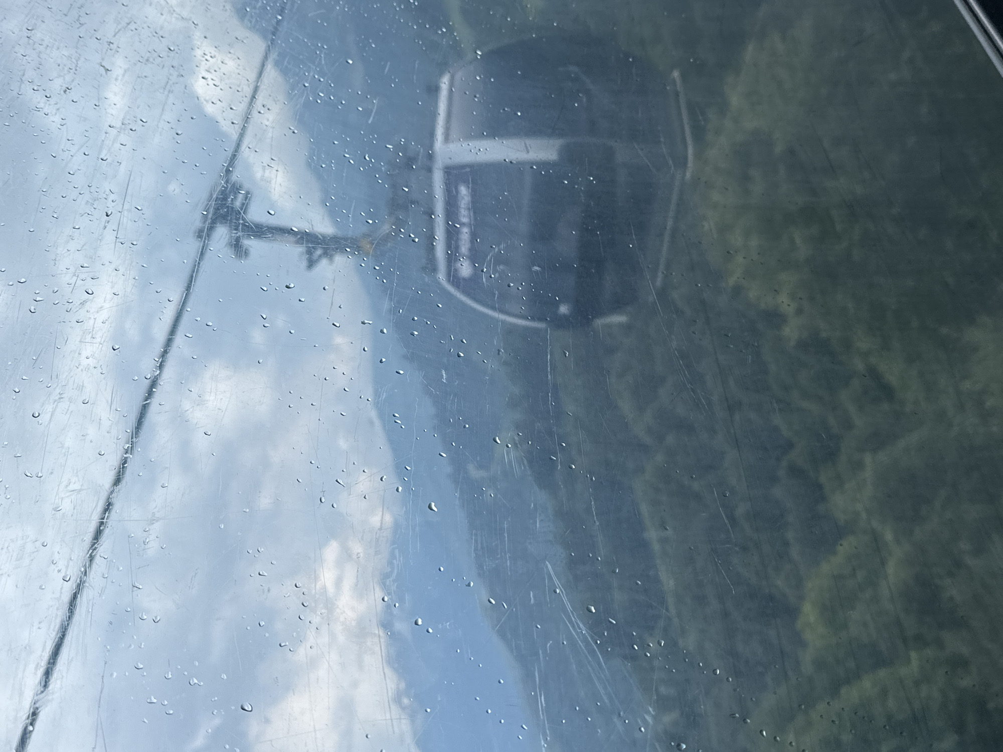 The image, taken from inside a cable car in Estosadok, Russia, shows a foggy, overcast day with rain on the window. The cable car itself is visible in the foreground, a dark, rounded cabin with white accents and writing on the front window. Through the rain-streaked glass, a cable car tower is visible in the distance, silhouetted against the hazy sky. The landscape below is mountainous and covered in lush, green trees. The overall atmosphere is muted and somewhat melancholic, due to the weather conditions.
