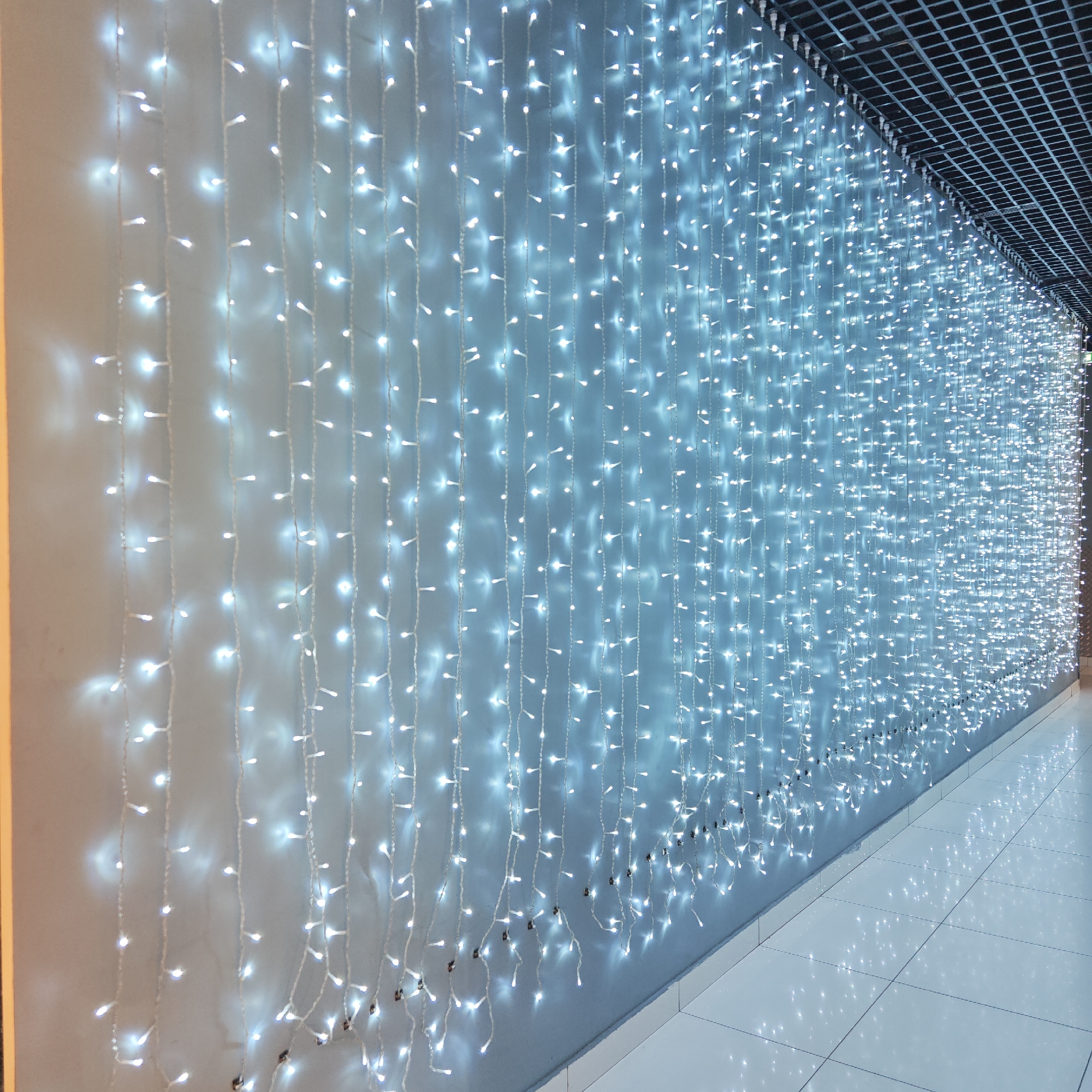 The image shows a wall adorned with strings of white LED lights, possibly in a commercial setting. The lights are densely packed, creating a luminous effect along the wall. The location appears to be an interior corridor with tiled flooring and a dark, possibly metal, grid ceiling. The photo's origin is Taganrog City District, Russia. There are no people visible in the image.