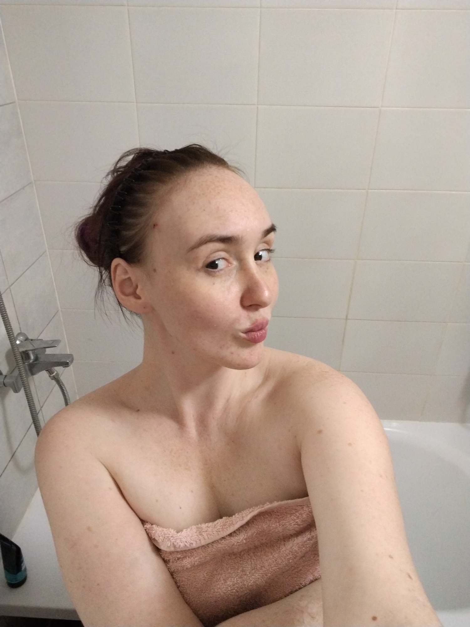 A young adult female is captured in a close-up portrait, presumably a selfie, within a bathroom setting in Izhevsk, Russia. She has fair skin with numerous freckles visible across her face, forehead, shoulders, and arms. Her dark hair is styled in a bun, revealing a subtle purple or reddish hue in places. She is looking directly at the camera over her right shoulder, her head slightly tilted, and her lips are pursed in a playful or pouting expression.
She is wrapped in a light peach or pink towel, which covers her chest and torso. The background consists of light-colored, rectangular wall tiles, suggesting a typical bathroom environment. To her left, a chrome shower fixture with a flexible hose and shower head is mounted on the tiled wall. The white rim of a bathtub is visible along the bottom and right edges of the frame. A dark bottle with a teal label, possibly a cosmetic or bath product, rests on the tub's edge in the lower-left corner. No text is visible on any objects. The image conveys a casual and intimate moment, likely taken after a bath or shower.
