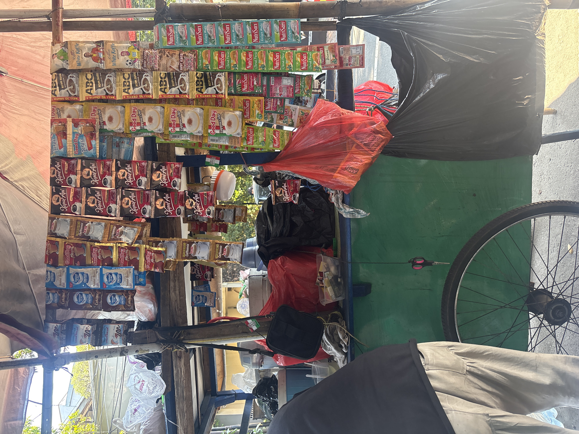 The image depicts a street vendor's stall in South Jakarta, Indonesia, displaying a variety of packaged coffee and powdered drink products. The stall is set up on a wooden cart with a green painted body, and a bicycle wheel is visible on the left side. The products are hung in rows from the overhead canopy structure, creating a colorful display. Among the visible brands are "Kapal Api," "Good Day," "ABC Kopi Susu," and "NutriSari." There are also some loose items like plastic bags, a black shoulder bag, and a clear container with cigarettes. A person's legs and lower torso, wearing light-colored pants, are visible in the foreground on the left. The background shows glimpses of buildings and greenery, typical of an urban environment. The overall scene suggests a typical roadside commerce setup in Indonesia.