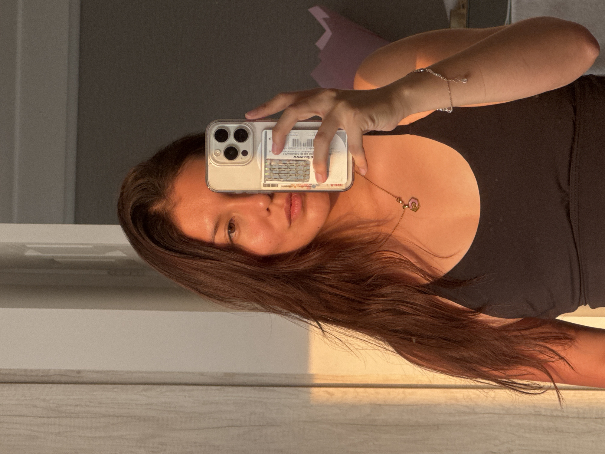 The image captures a mirror selfie taken by a person, presented at a 90-degree counter-clockwise rotation, likely in San Francisco Ocotlán, Mexico. The subject, with long, flowing dark brown hair, is wearing a dark top, a delicate silver bracelet, and a gold-toned necklace featuring a small, pink or purple faceted pendant.
She holds a light-colored smartphone, possibly an iPhone with a triple camera system, in a clear case, which partially obscures her face. Within the phone case, several items are visible, including cards or slips of paper. Notable text includes "ebiu.www," Cyrillic letters "ИЯЕ," and dates "01.07.2023" and "01.07.2024," along with a barcode and what appears to be a metallic chip card. Her brown eyes are visible, and her lips are slightly parted in a neutral expression.
The reflected background reveals an indoor setting with a dark grey wall meeting a lighter, possibly white, wall or door frame. A wooden surface, such as a floor or countertop, is visible at the bottom of the reflection. The scene is illuminated by warm, golden light, indicative of either sunrise or sunset, casting a soft glow on the subject's skin and hair. No explicit location cues specific to San Francisco Ocotlán are visible within the image content.
