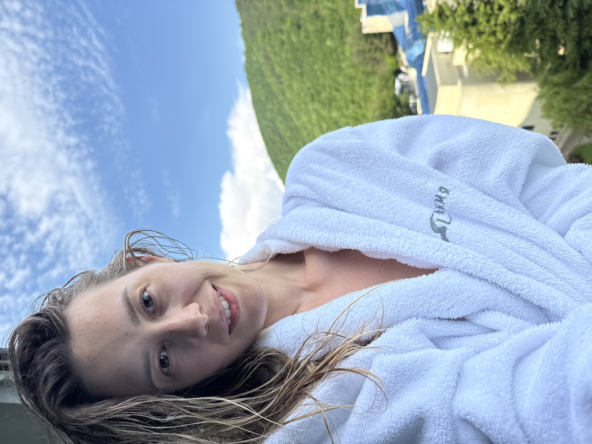 The image shows a woman in a white bathrobe standing on a balcony in Nebug, Russia. She has wet hair and is smiling at the camera. In the background, there is a lush green mountain and a clear blue sky with scattered clouds. A building complex is visible at the base of the mountain. The woman is the primary subject, positioned in the foreground, and the natural landscape forms the background.