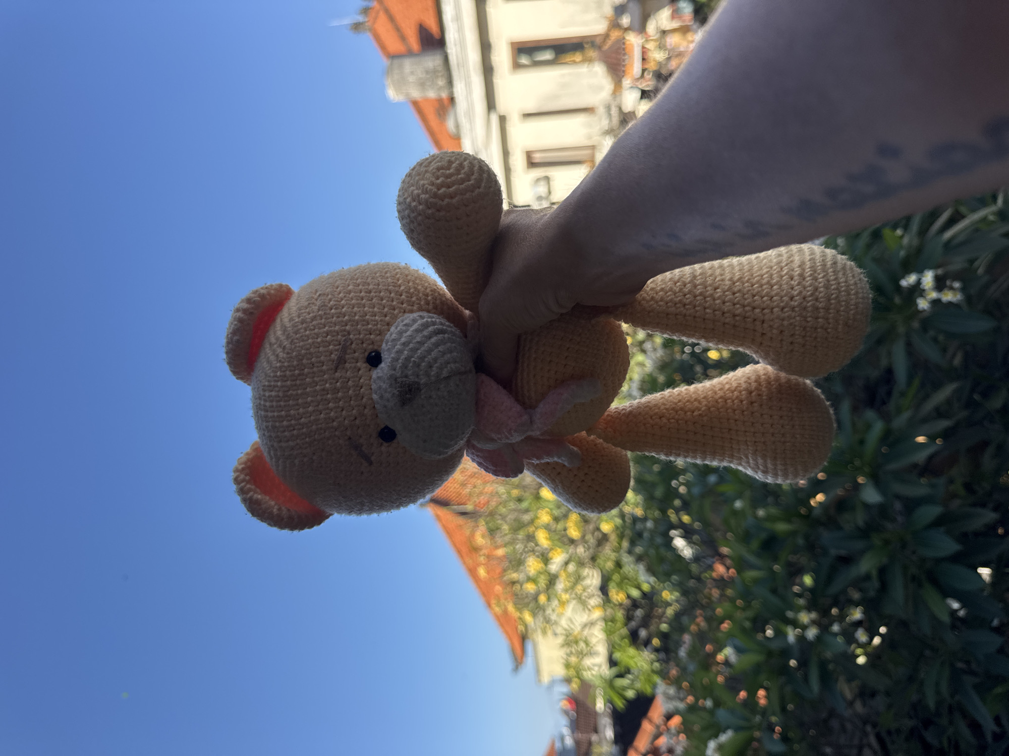 A person's hand holds a light brown crocheted teddy bear with a pink scarf against a bright blue sky. Behind the bear and hand, out-of-focus elements suggest a cityscape with red-tiled roofs and lush green foliage. The scene appears to be in Kuta, Indonesia, given the architectural style of the buildings and the tropical vegetation. The teddy bear is positioned in the foreground, centrally located, with its legs dangling. The person's arm, visible from the wrist up, is tattooed. The lighting indicates it is daytime.