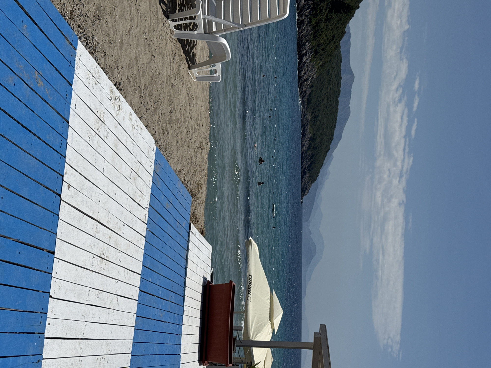 A beach scene with a wooden walkway painted with blue and white stripes in the foreground. To the left, a white lounge chair sits on the sand. The ocean is calm with several people swimming in it. In the distance, lush green hills and mountains rise against a blue sky with scattered clouds. Two large beige umbrellas with "Tuborg" branding are visible near the water's edge, with a red planter box beneath them. The scene appears to be a resort or a public beach.