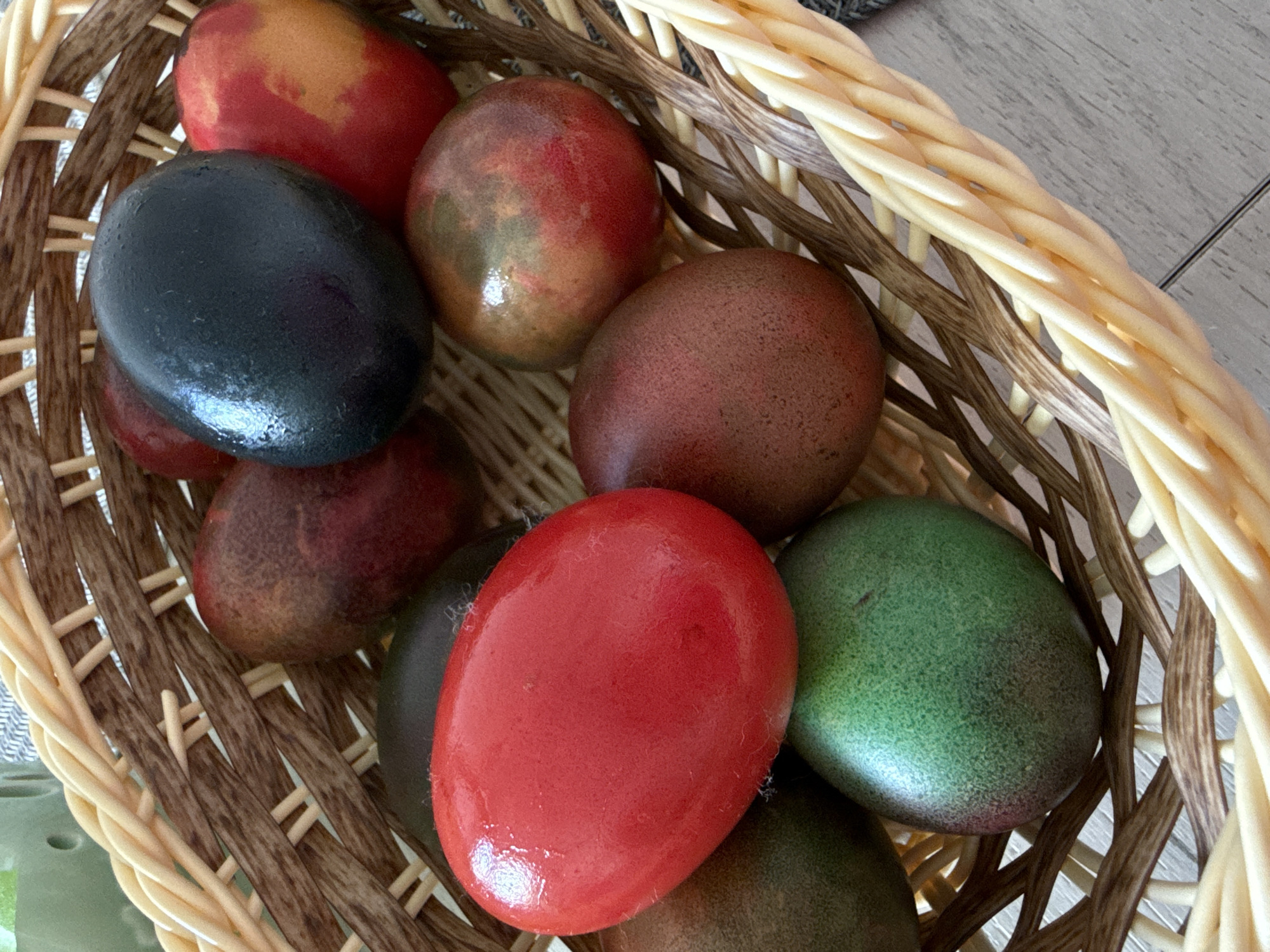 A close-up, high-angle shot shows a wicker basket filled with colorful, egg-shaped objects. These objects appear to be painted or dyed in various colors, including red, black, green, and shades of brown and red-brown, some with mottled patterns. The lighting is diffused, creating highlights on the smooth surfaces of the objects. The basket is constructed of light and dark tan woven material. The background is out of focus, but appears to be a patterned surface, possibly a tablecloth or flooring. The scene is likely indoors. There is no indication of the city of Sofia or Bulgaria.