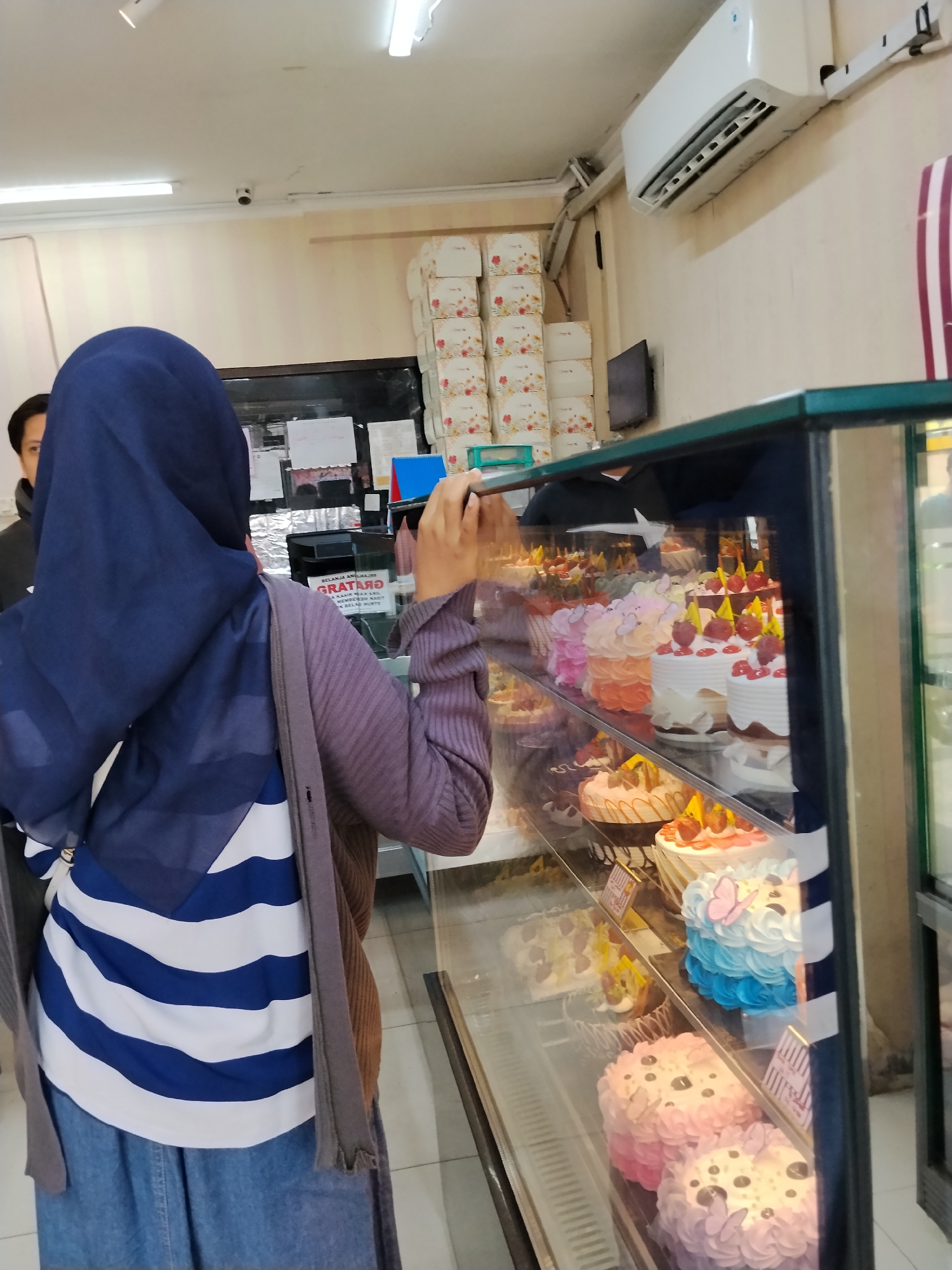 In Ciruas, Indonesia, a person wearing a dark blue hijab and a blue and white striped shirt is standing in front of a bakery display case filled with various cakes. They are wearing a purple cardigan and a denim skirt. Their right hand is raised, holding a phone and taking a picture of the cakes. Another person is partially visible behind them. The display case shows several tiers of cakes with different frosting colors and decorations, including some with fruit and edible flowers. Boxes of what appear to be pastries or gift items are stacked on shelves behind the display. An air conditioning unit is mounted on the wall above the display. The lighting suggests an indoor environment, likely during the daytime.