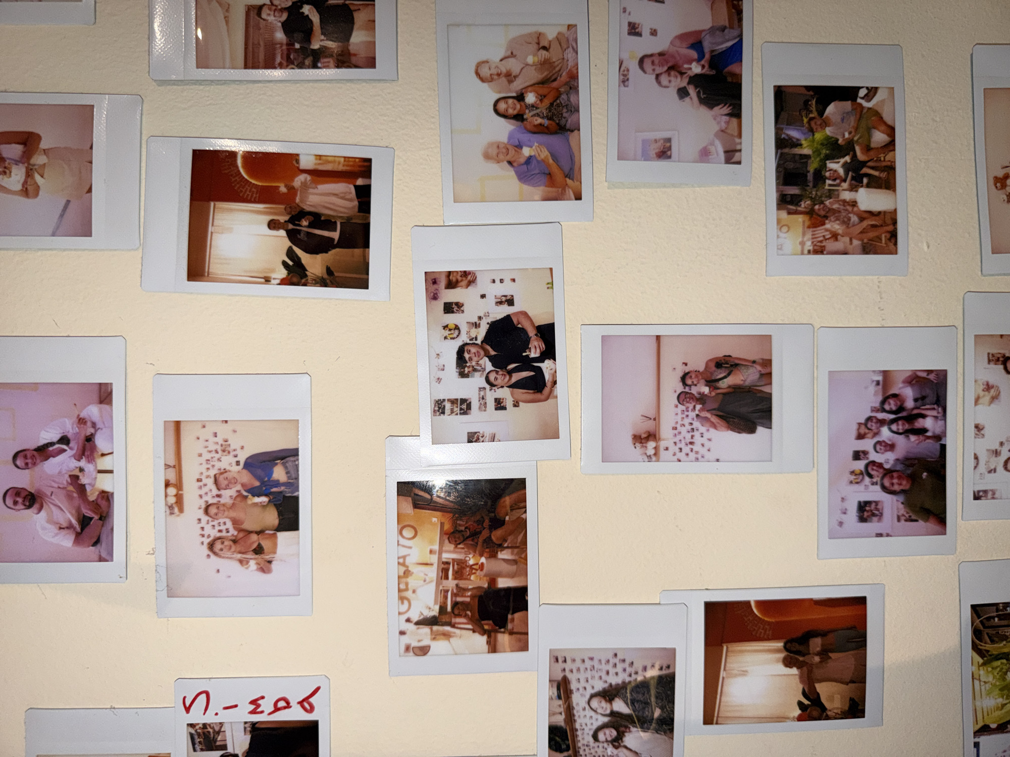 The image shows a collection of instant photos, likely Polaroids, affixed to a light yellow wall. The photos depict various subjects: groups of people posing indoors and outdoors, scenes from cafes and restaurants (one with "GELATO" visible), and images of people in indoor settings, some with walls covered in more photos. A piece of red writing appears near the bottom of the frame. The location, North Kuta, Indonesia, is not discernible within the photographic evidence of the scene.