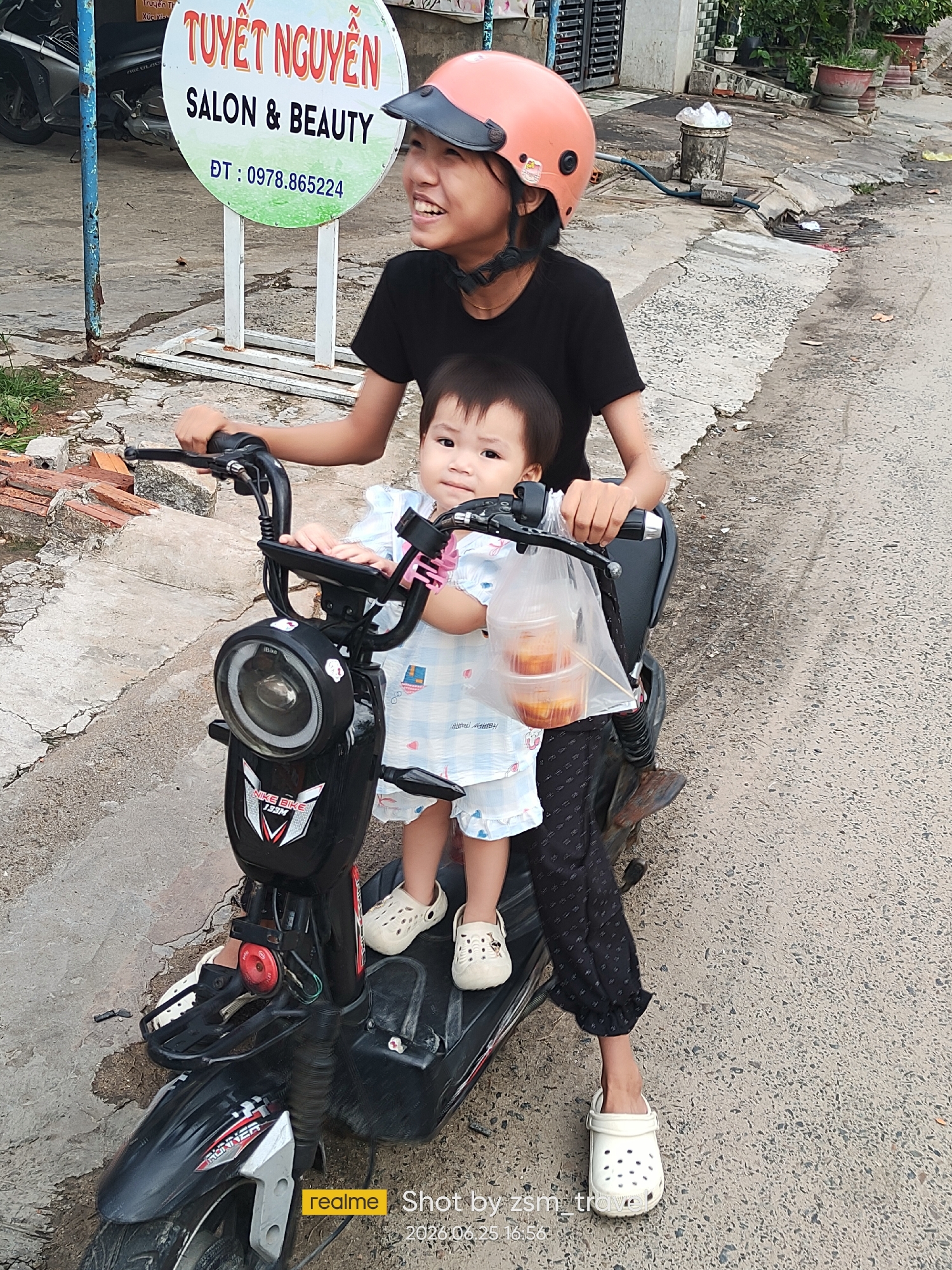 Two young females, one older, are seated on an electric scooter in Phu Thuy Ward, Vietnam. The elder, wearing a black t-shirt and an orange helmet, is driving. The younger child, in a blue and white checkered outfit, sits in front. A plastic bag containing two small containers of food is hanging from the handlebars. The scooter is black with red and white accents. They are on a paved road with a sidewalk and some greenery visible in the background, along with a circular sign indicating "TUYET NGUYEN SALON & BEAUTY" with a phone number. The timestamp "2023.06.25 16:56" is visible in the bottom right corner, along with "realme Shot by zsm_trove".