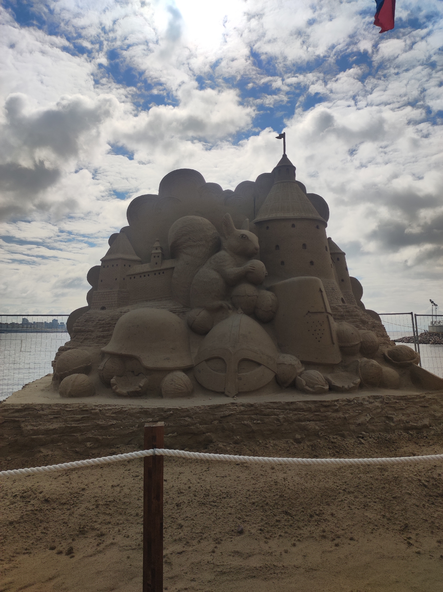 A massive sand sculpture depicting a fantastical castle is the central subject of the image. The sculpture features multiple turrets, walls, and even a squirrel perched on top, holding what appears to be a nut or a round object. In front of the castle are various objects carved from sand, including what look like helmets and shields, suggesting a historical or mythological theme. The sand sculpture is situated on a sandy beach, with a body of water, likely the Gulf of Finland, visible in the background. A wire fence separates the sculpture from the water and the surrounding area. In the distance, across the water, a city skyline can be seen, which, given the context of Saint Petersburg, Russia, likely represents the city's silhouette. The sky above is filled with dramatic clouds, with patches of sunlight breaking through, indicating a partly cloudy day. A red and blue flag is flying in the upper right corner of the image. A rope barrier with wooden posts is in the foreground, indicating a viewing area for visitors. The overall impression is of a public art installation on a beach.