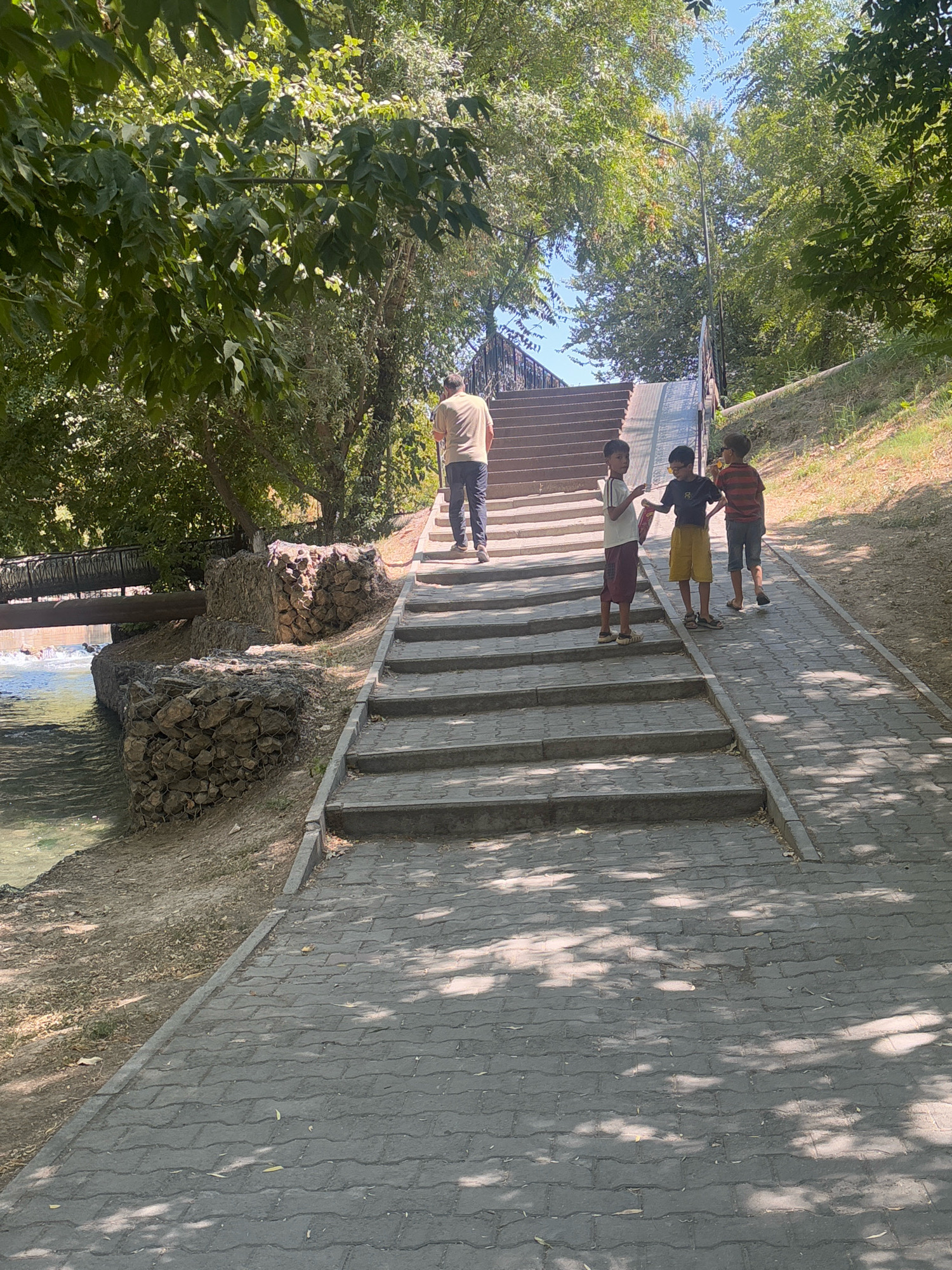 A group of four individuals, including one adult male and three young boys, are present in a park-like setting. The adult male is walking up a set of stone steps, while the three boys are standing together on a paved pathway adjacent to the steps. One boy is wearing a striped shirt, another a dark t-shirt and yellow shorts, and the third is in a white t-shirt and maroon shorts. The environment features lush green trees providing shade, a body of water with a bridge crossing it to the left, and gabion walls constructed with rocks along the water's edge. The pathway and steps are made of interlocking pavers and stone, respectively. The scene suggests a casual outdoor activity on a sunny day. This scene is located at the Shymkent Zoo in Shymkent, Kazakhstan, specifically near the bear enclosures.