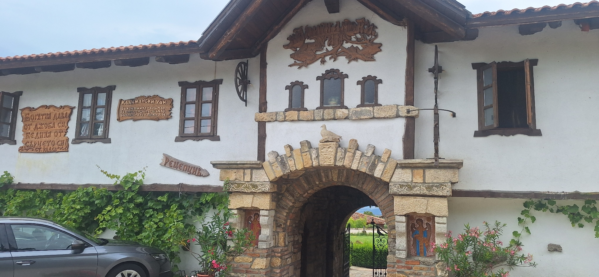 The image shows a white-plastered building with a tiled roof and wooden window frames. The building features several signs with Cyrillic writing. Above an arched stone entryway, a wooden carving depicts animals, possibly deer, on a branch. A stone pigeon statue sits atop the archway. The entryway itself is constructed from rough-hewn stones. To the left of the entryway, a gray car is parked on a paved surface partially covered by green ivy. To the right of the entryway, an image of an angel is visible on the stone pillar. Greenery, including vines and flowering plants, surrounds the lower part of the building. The overall scene suggests a historical or cultural site, possibly an inn or a monument, in the town of Kalimantsi, Bulgaria.