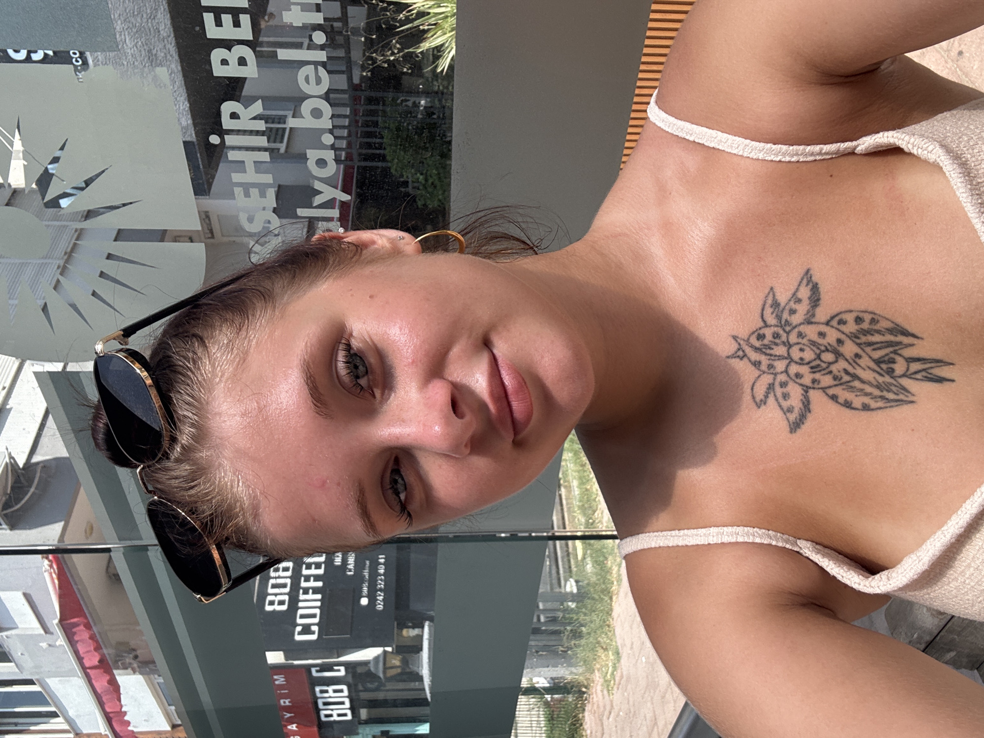 The image shows a young woman with her hair pulled up in a bun, wearing sunglasses on top of her head. She has a tattoo on her chest and is wearing a light-colored tank top. She is outdoors, and in the background, there are signs for "808 COIFFEUR" and "ŞEHİR BEL." which suggests the location is in Antalya, Turkey. The setting appears to be a street or a cafe with buildings and vegetation visible.