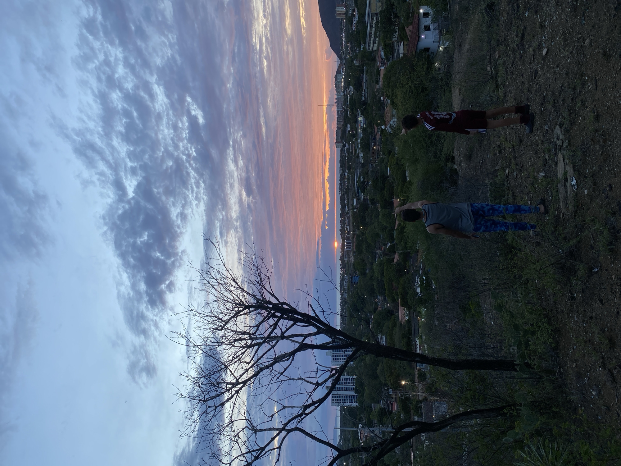 Two young males stand on an elevated dirt and scrub-covered hill overlooking a coastal city during a sunset. The male on the left wears a gray tank top and patterned blue and white pants. He is facing away from the camera, pointing his left arm upwards towards the sky. The male on the right wears a maroon and white athletic jersey and maroon shorts. He is facing away from the camera, pointing his right arm towards the cityscape. The sky is filled with scattered clouds colored in hues of blue, pink, and orange, indicating the sun is setting on the horizon over the ocean. In the mid-ground, buildings, trees, and streetlights form the cityscape. In the background, a mountain range is visible to the right. A bare tree stands in the foreground to the left of the frame. The scene suggests a quiet, observational moment at dusk in Juan Griego, Venezuela.
