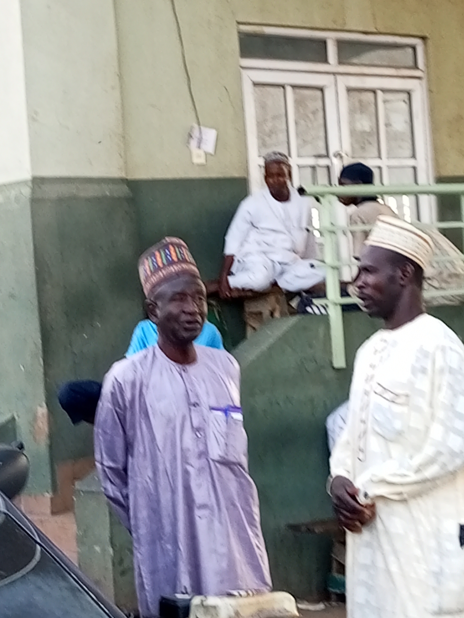 In the city of Gombe, Nigeria, an outdoor scene depicts several men gathered near a building with a white-framed window. In the foreground, two men are prominent. The man on the left wears a purple thobe and a patterned kufi, while the man on the right is dressed in a white thobe and a white and black striped kufi. Behind them, other men are visible, one sitting and wearing a white garment and a white kufi, and another in the background wearing a dark blue kufi. The environment appears to be a courtyard or entrance area with a weathered green wall and hints of concrete structures. The lighting suggests it is daytime.