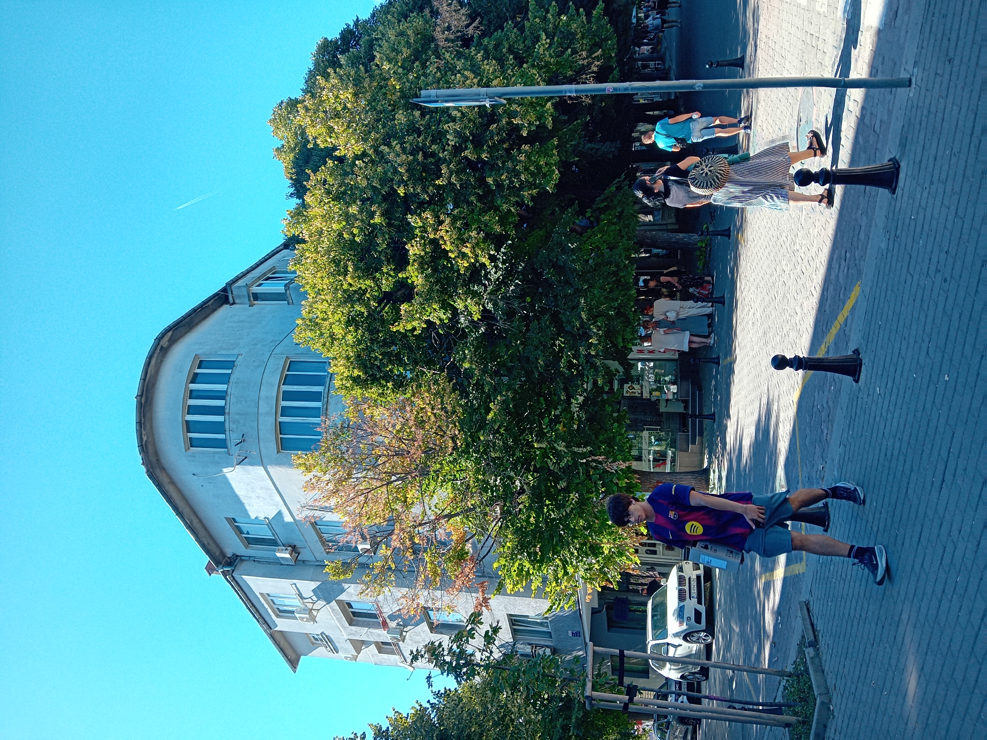 In Varna, Bulgaria, on a sunny day, a young man with glasses, wearing a purple and red t-shirt with a blue collar and a Spotify logo, and grey shorts, walks on a brick-paved sidewalk carrying a light brown box and his phone. He is in the foreground, walking from left to right. Behind him, a woman with dark hair, wearing a black top and a black and white striped skirt, walks while talking on her phone. She has a round, straw-like bag on her back. Further behind, other people are seen walking on the sidewalk. To the left of the frame, a white SUV is parked, and trees with green and yellow leaves line the street. A curved, light-colored building with multiple windows is visible in the background. The sky is clear and blue, with a faint airplane contrail visible. The scene appears to be a street in a city with a mix of residential and commercial buildings, with pedestrian activity.