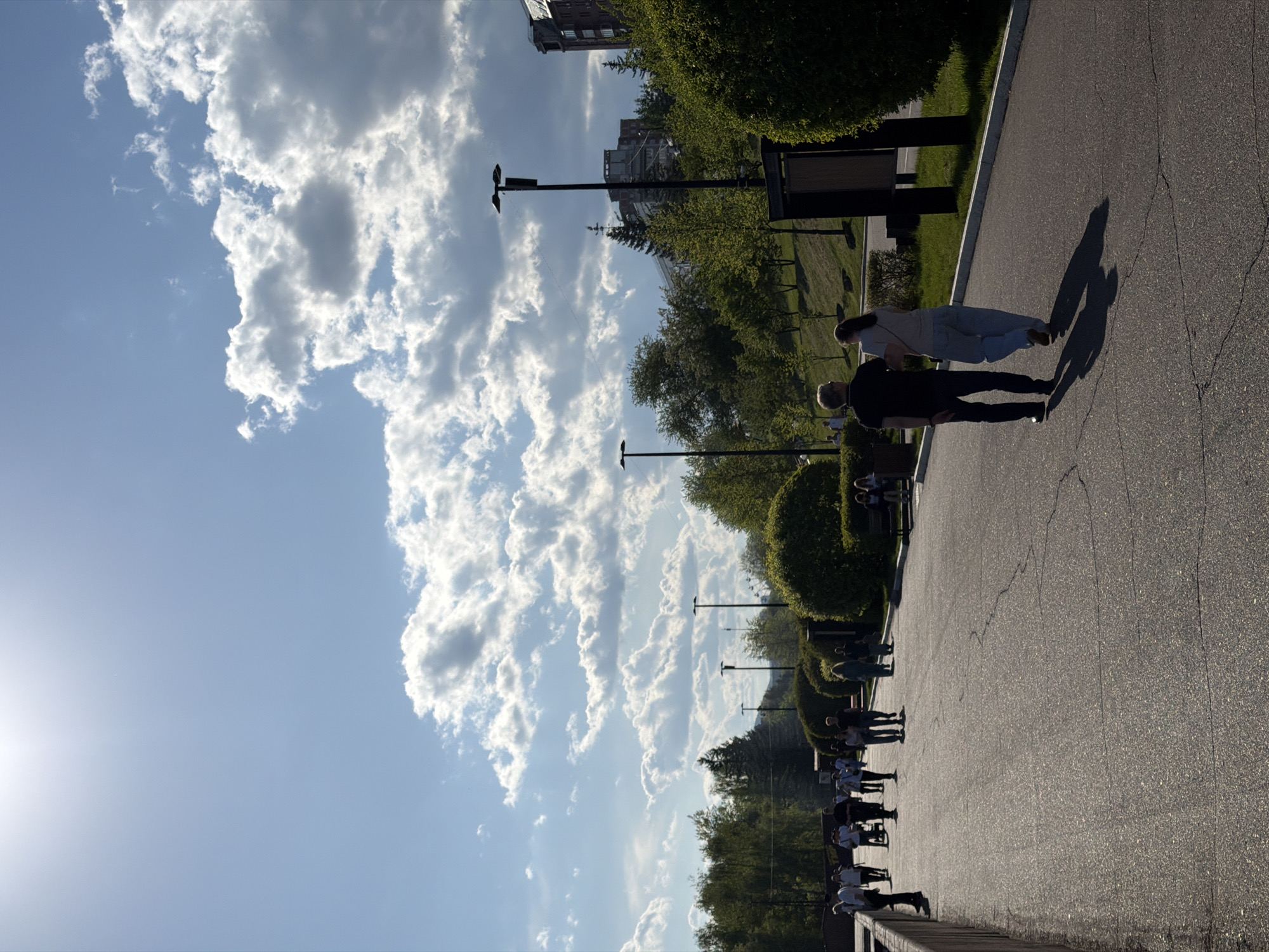 A landscape-oriented, eye-level photograph depicts a sunny scene along a road, likely a pathway, in Krasnoyarsk, Russia. A bright sun shines from the left side of the image, illuminating fluffy white clouds that dominate the left side of the frame.
The paved road occupies the right side of the image. Several people are walking along the road. A couple is visible near the middle, while a larger group, is observed at the bottom.
Trees and manicured shrubbery line the left side of the pathway. Lamp posts are evenly spaced along the path, extending towards the top-left corner of the image. In the distant background, a building structure is visible. The overall composition suggests a warm day in a well-maintained urban park or similar public space in Krasnoyarsk, Russia.