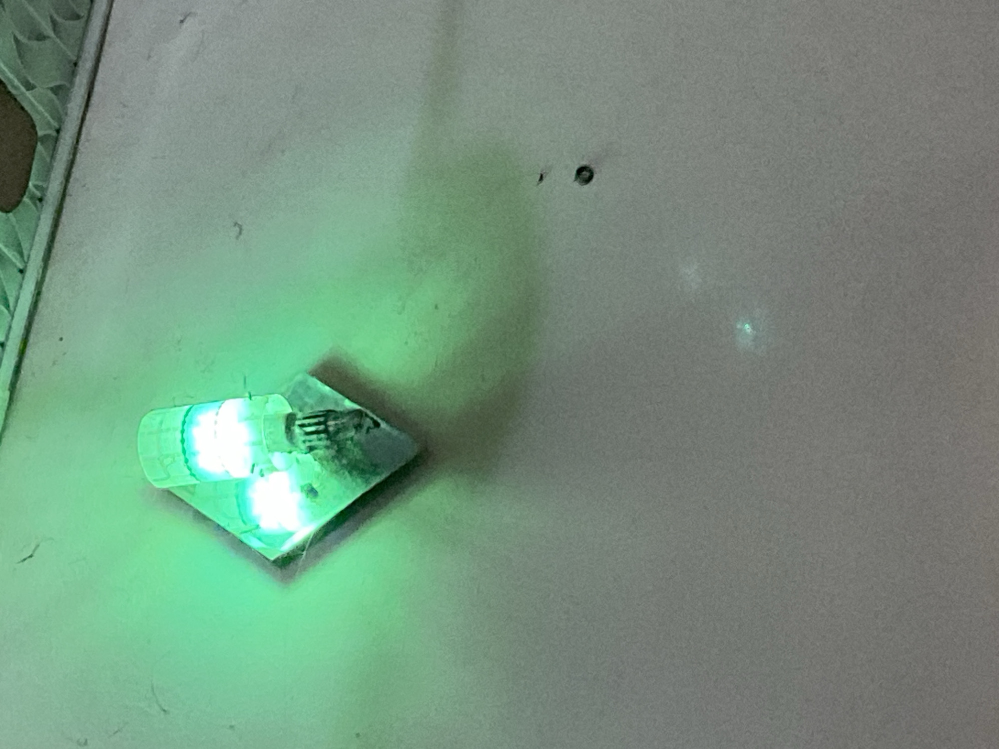 A green LED light fixture is mounted on a light-colored wall in Mararaba, Nigeria. The fixture is diamond-shaped and has a clear, ribbed cover over the light source, which emits a bright green glow. The light casts a soft shadow on the wall. The wall itself appears textured and shows some minor imperfections. The top of the image shows a glimpse of a ceiling or overhead structure.