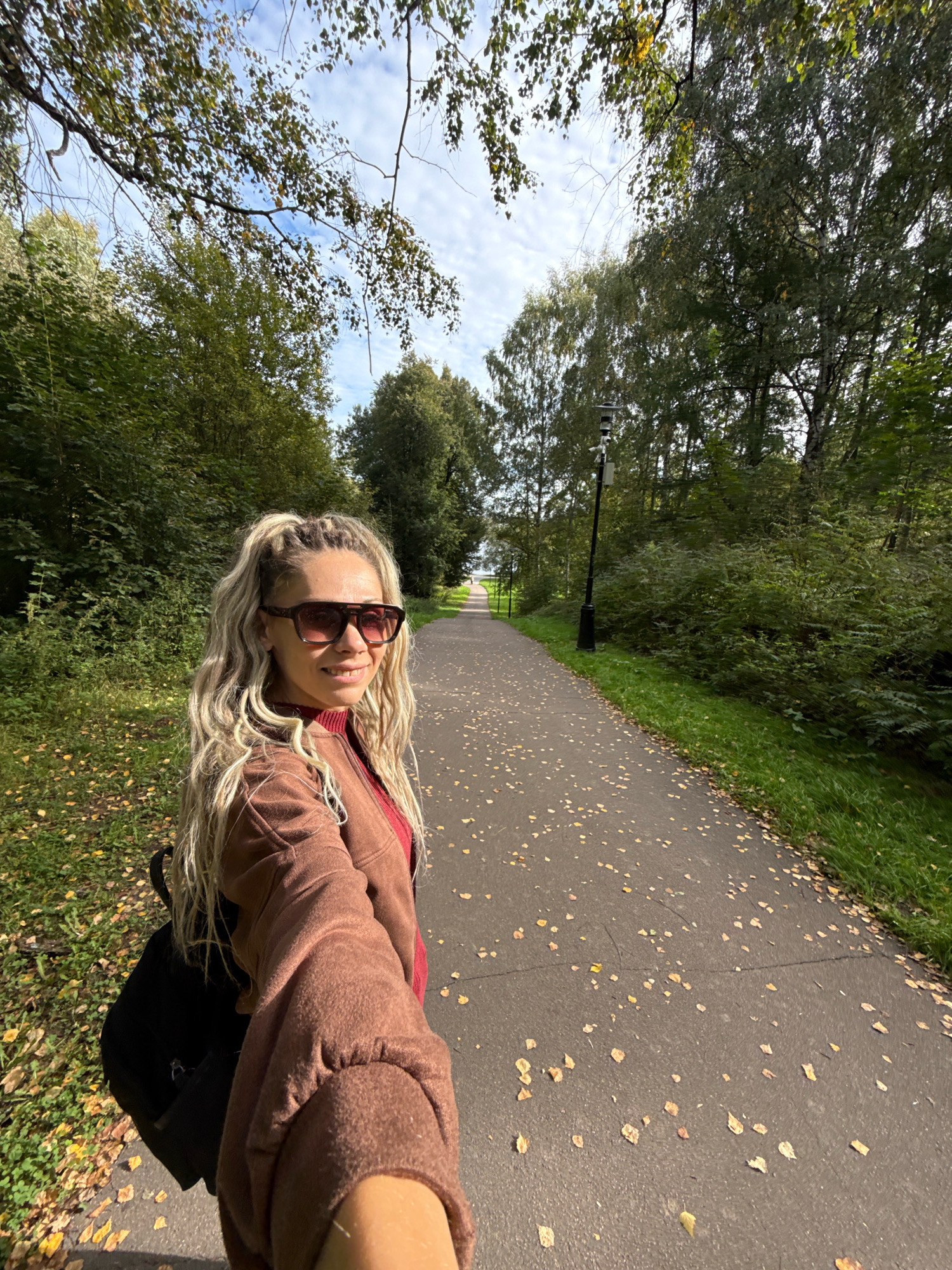A woman with blonde hair in a high ponytail, wearing large sunglasses, a red shirt, and a brown jacket, smiles at the camera in a selfie. She is walking on a paved path lined with trees and grass. The path is covered with fallen yellow leaves, indicating it is autumn. A black backpack is visible on her back. In the background, a lamppost stands on the side of the path. The location is identified as Rusanovo, Russia, suggesting a park or wooded area within the city. The sky is partly cloudy.