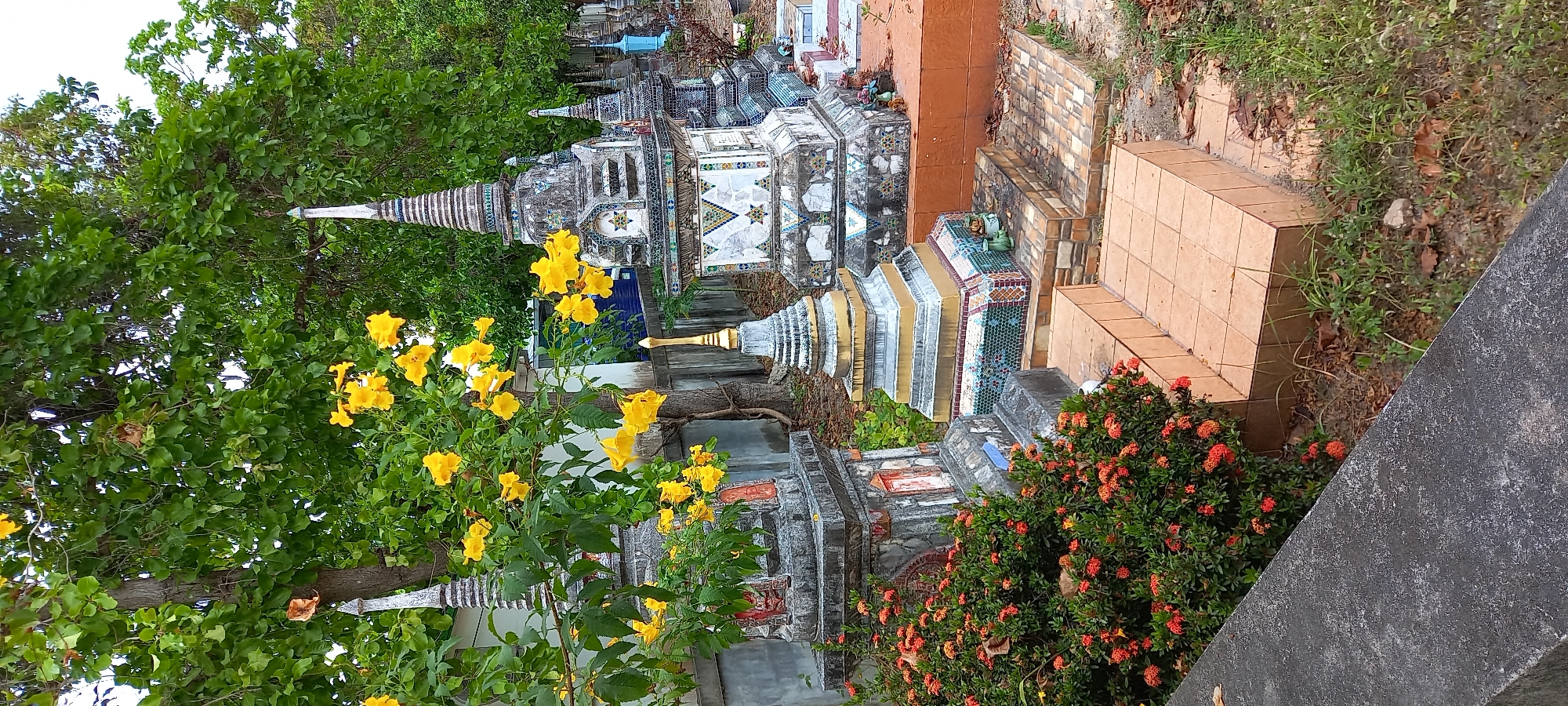 The image shows a section of a graveyard in Moo 4, Thailand. The graves are ornate, featuring tiered, pagoda-like structures decorated with colorful tiles and geometric patterns. Lush greenery surrounds the graves, including a bush with vibrant yellow flowers and another with clusters of small red flowers. Some graves are constructed of orange-colored bricks or tiles. A gray, concrete edge runs along the lower right of the image.