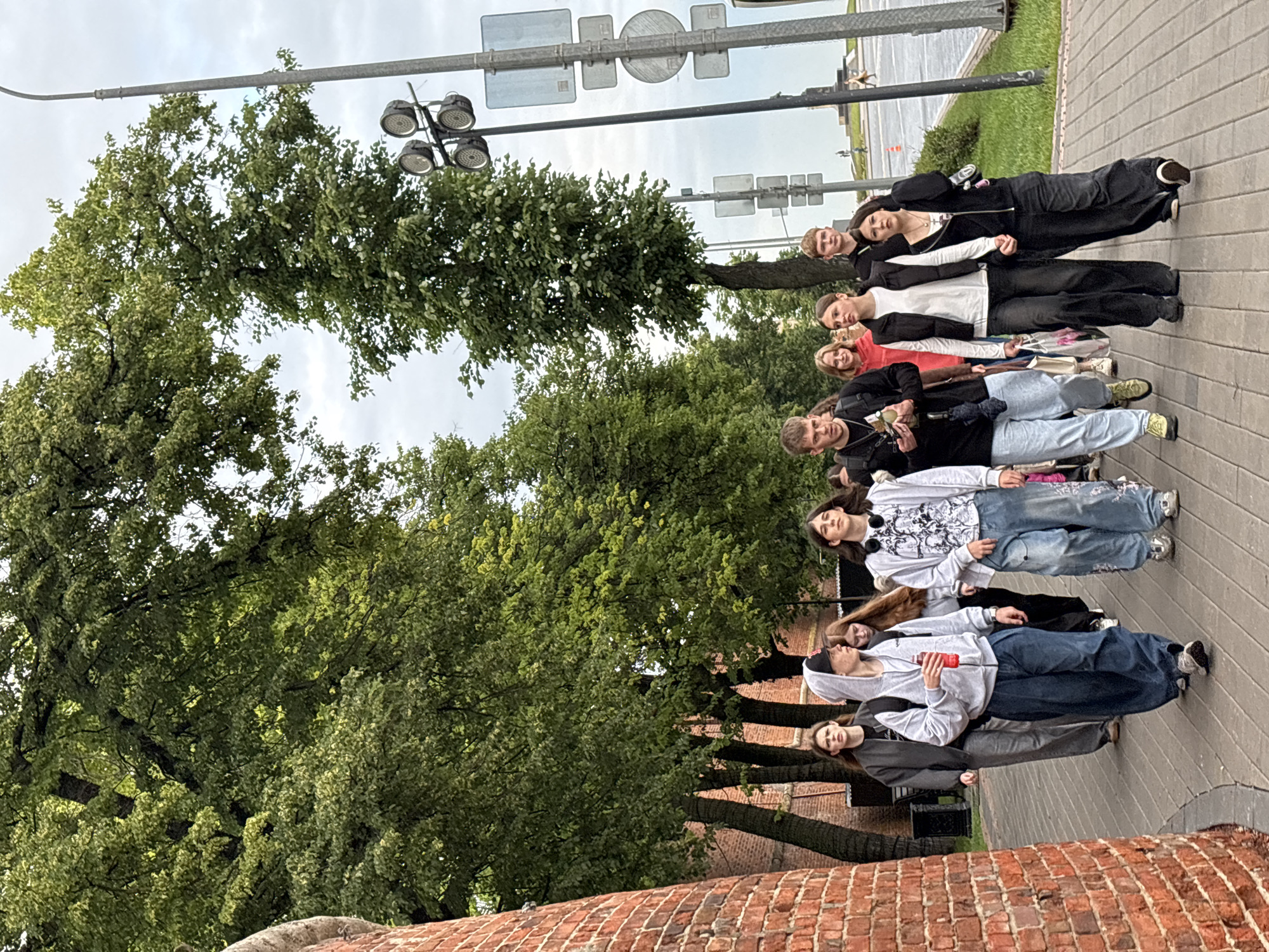 A group of eight young people, appearing to be in their late teens or early twenties, are walking down a paved sidewalk. The group is diverse in gender and is dressed casually in streetwear, including hoodies, jeans, and sneakers. Some individuals are carrying backpacks or bags. They are walking in the same direction, and their expressions suggest they are engaged in conversation or are simply walking together.
The environment appears to be an urban setting, possibly a park or a boulevard, with a brick wall on the left and a line of trees with lush green foliage on the right. In the background, a lamppost with multiple lights and traffic signals is visible, along with street signs. The sky is overcast, suggesting a cloudy day. In the far background, there is a glimpse of a distant monument or statue. The overall scene suggests a casual outing or exploration of a public space. Based on the context provided by the prompt indicating the location as Nizhny Novgorod, Russia, this scene is likely situated in a prominent public area within the city, possibly near a historical site or a recreational zone.
