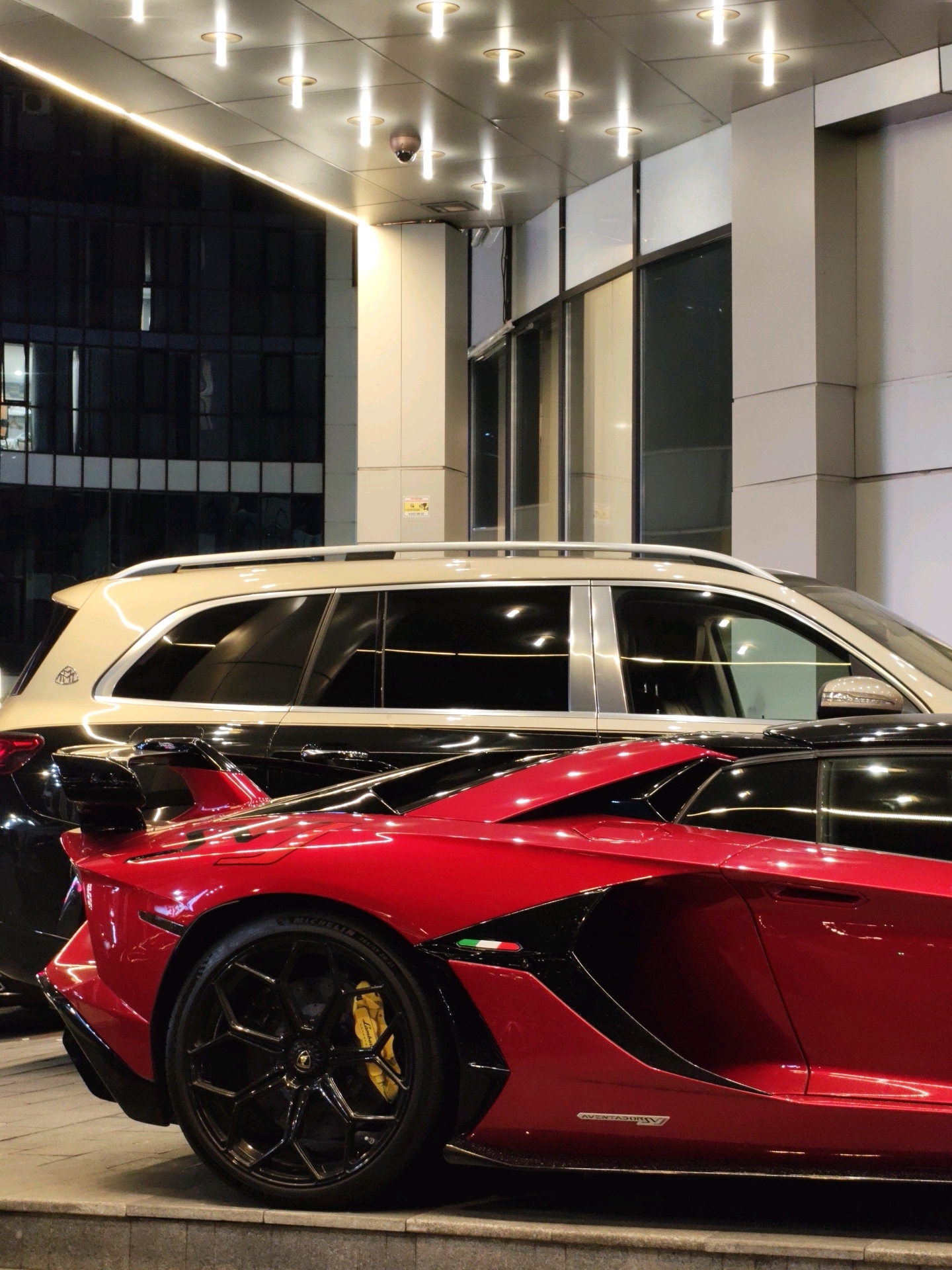 A red Lamborghini Aventador is parked on a tiered stone surface in front of a modern building. The car is angled, showing its rear quarter panel, with a prominent black wing and a black wheel with yellow brake calipers. Behind the Lamborghini, a black SUV with a cream-colored roofline, identified as a Maybach, is partially visible. The building's facade features large windows and a metallic overhang with recessed lighting. The scene is lit by artificial lights, creating reflections on the vehicles and the building's surfaces. The image appears to have been taken at night. The location is the city of Batumi, Georgia, likely near a high-end establishment or residence. There are no people visible in the image.