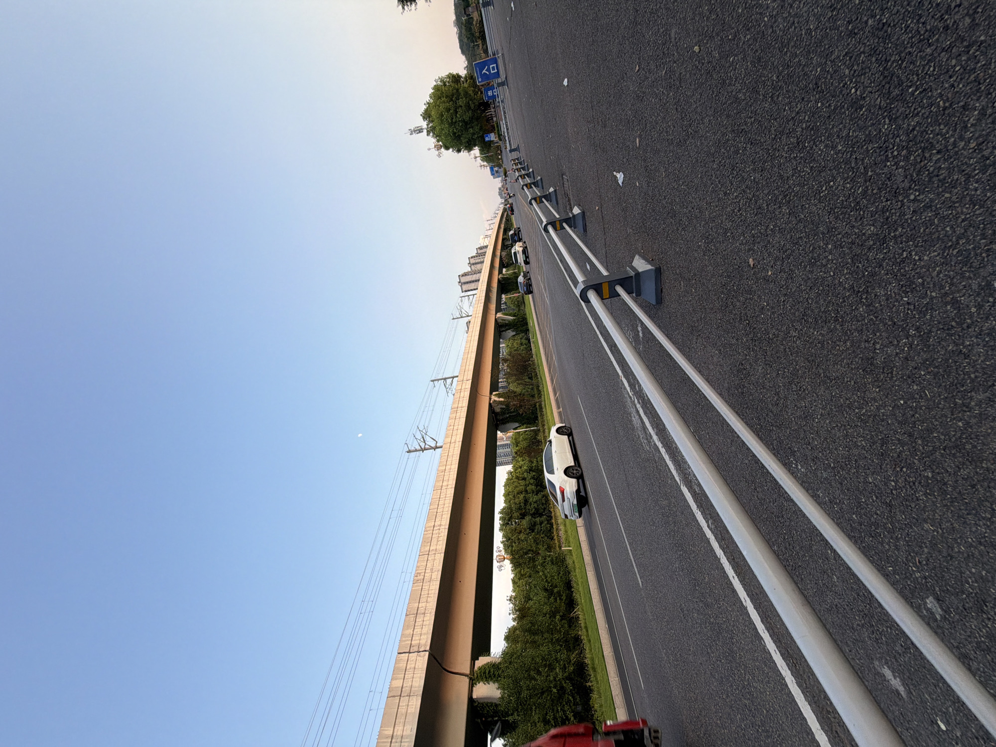 The image captures a scene in Jiangjin District, Chongqing, China, likely during the late afternoon or early evening. The primary focus is a multi-lane road with a light rail track elevated above it. The sky is a clear, pale blue with a hint of a crescent moon visible.
On the road, several vehicles are present. A white sedan is in the left lane, heading away from the viewer. Further down the road, other vehicles are visible, including a black sedan and a motorcycle. The road surface is asphalt with white lane markings. Guardrails line the right side of the road, separating it from the shoulder and a grassy area.
The elevated structure is a concrete bridge supporting the light rail, with visible overhead power lines. It curves into the distance. Below the bridge, lush green trees and bushes form a dense line. In the background, several multi-story buildings suggest an urban or suburban setting.
To the right of the road, a sign with Chinese characters, possibly indicating an entrance or exit, is visible. A lone tree stands near the sign. The ground in the foreground is also asphalt, with some scattered litter. A red vehicle is partially visible on the far left edge of the frame.