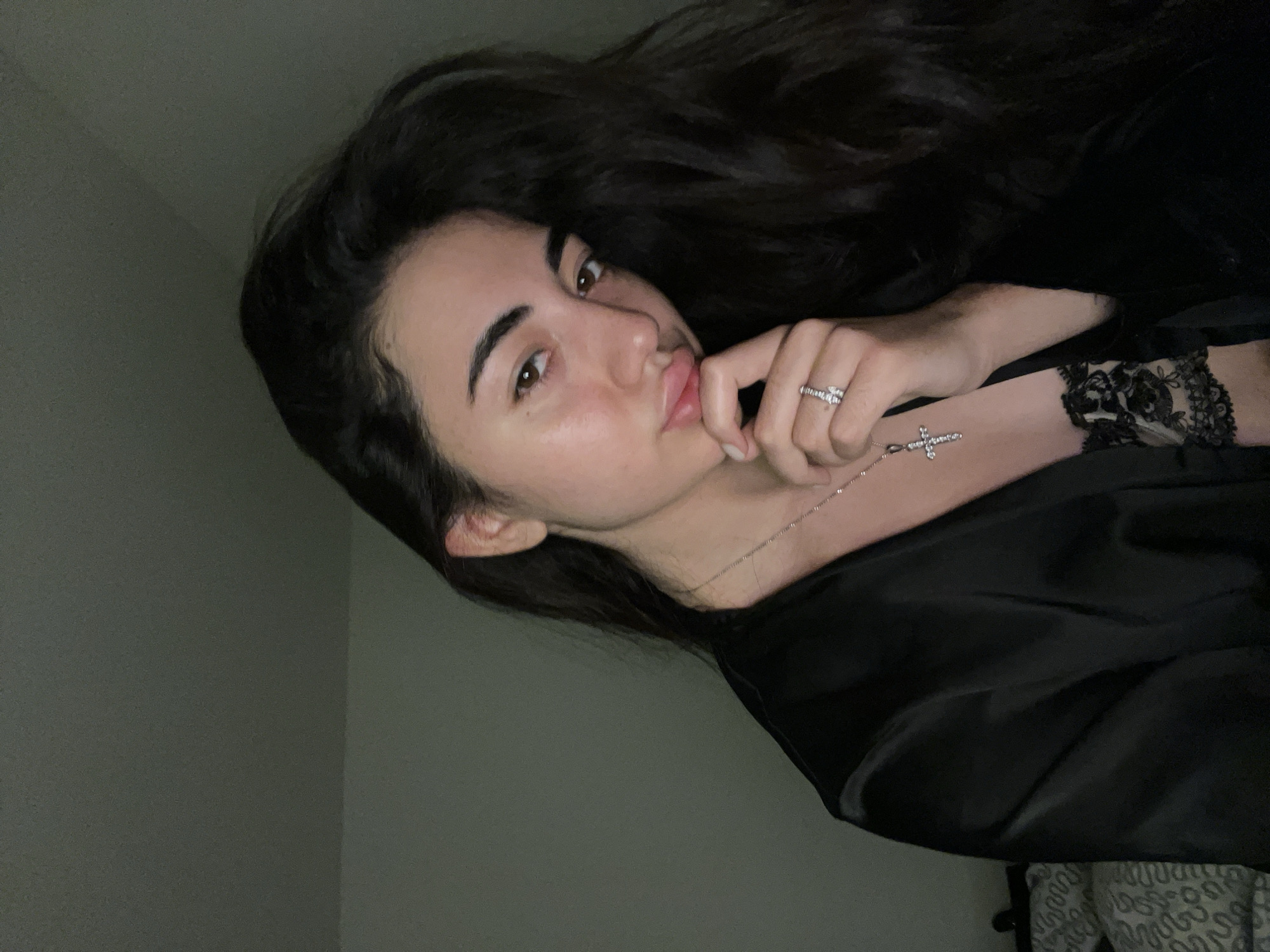 A young woman with long, dark hair is pictured from the chest up, looking to the viewer's right. She is wearing a black satin robe, with a glimpse of black lace peeking out from underneath. Around her neck, she wears a delicate silver chain with a cross pendant. On her left hand, she wears a ring on her ring finger. Her right hand is raised to her chin, with her index finger touching her lips. The background is a plain, dark wall, suggesting an indoor setting. The lighting is dim, with a spotlight on the woman's face. There is no indication of the location being in the city of Chotěšov, Czechia.