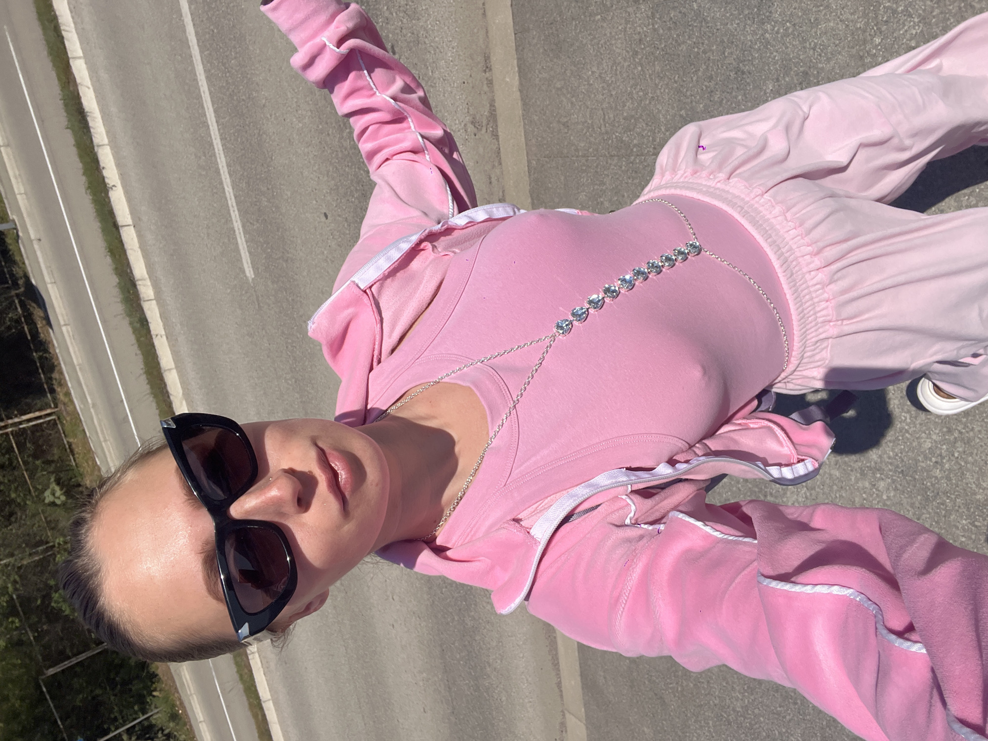 A high-angle selfie captures a person standing on an asphalt road, angled downwards and slightly tilted. The subject is wearing a two-piece pink velvet tracksuit with white piping and white zippers on the jacket, and a plain light pink top underneath. A silver-colored body chain with a line of large, round, clear gem-like stones adorns their torso over the pink top. A thin gold-colored chain necklace is also visible. The subject wears black, cat-eye style sunglasses and has their hair pulled back. Their expression is neutral.
The setting appears to be outdoors on a sunny day, evidenced by strong shadows and bright lighting. The ground is a grey asphalt surface with white road markings visible, suggesting a street or highway. To the left, there's a curb or barrier, beyond which some green foliage is visible. The pants of the tracksuit feature ribbed cuffs at the ankles and a gathered, elasticized waistband. Part of a white sneaker is visible on the subject's right foot. The location is contextualized as Boreti, Montenegro. There is no visible text in the image.
